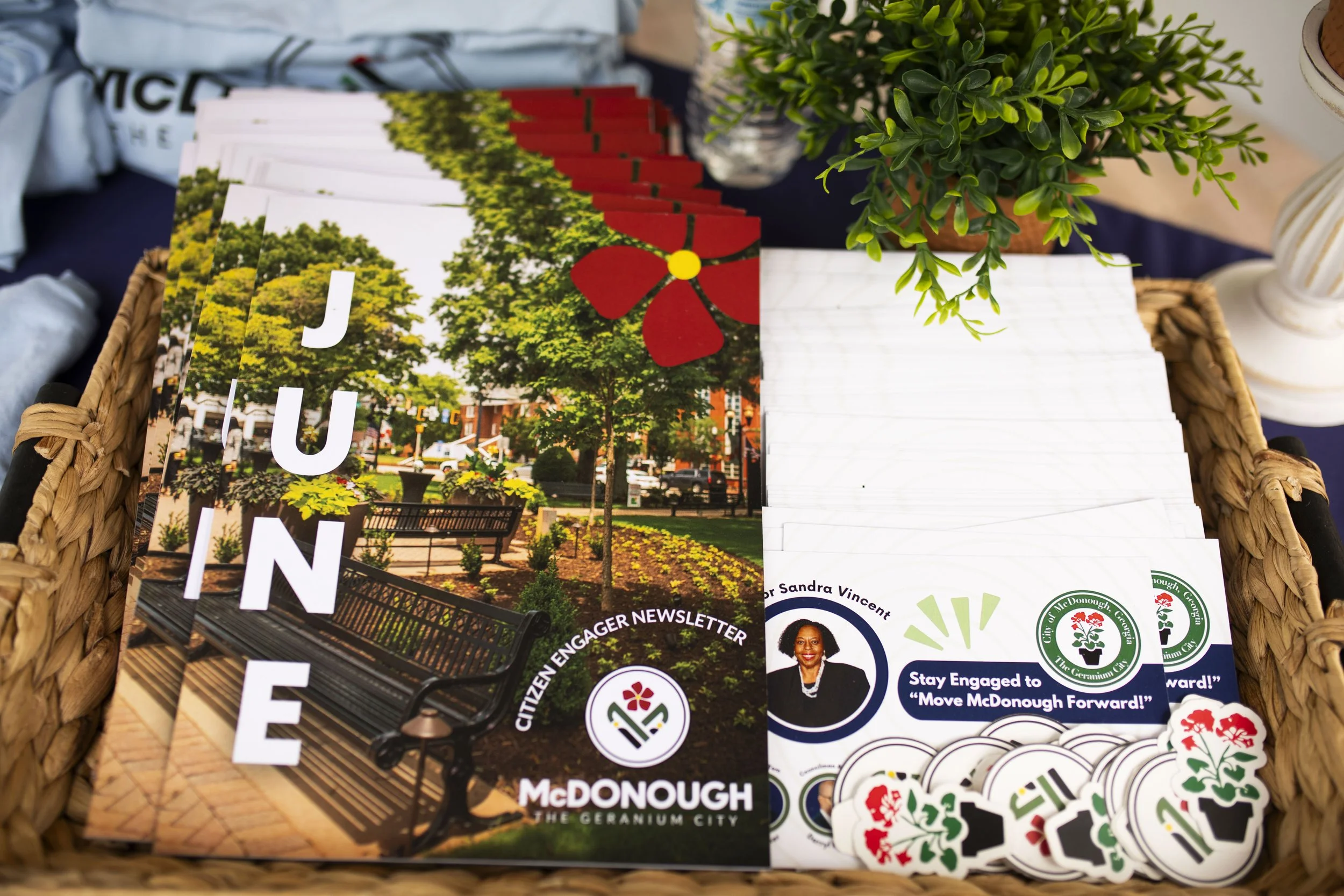 A basket containing flyers for McDonough, Georgia, featuring images of a park with trees and benches, alongside stickers with the city logo and a portrait of Sandra Vincent.