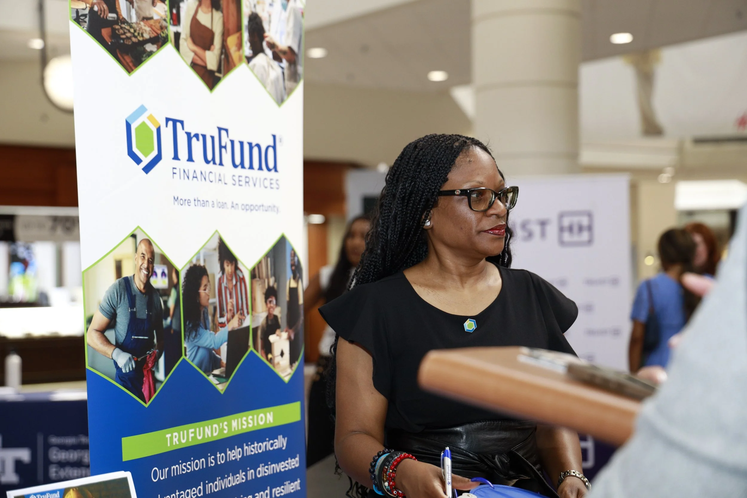 A woman with black hair, glasses, and a black dress standing at a booth with a Trufund Financial Services banner. The banner features images of people engaging in various activities and the company's mission statement.
