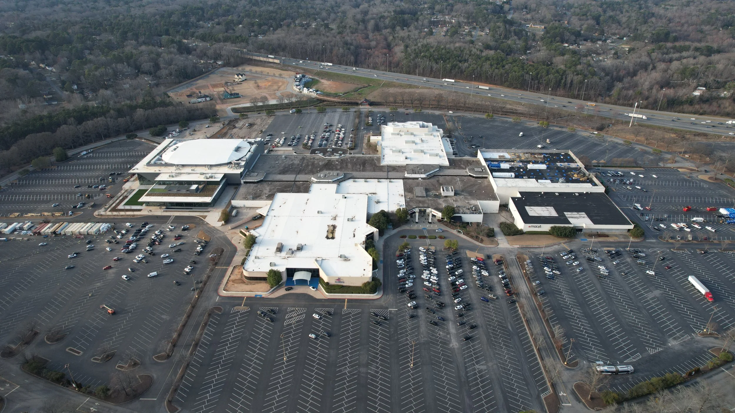 Aerial view of a large shopping mall with parking lots around it, some with cars parked, and a highway in the background surrounded by trees.