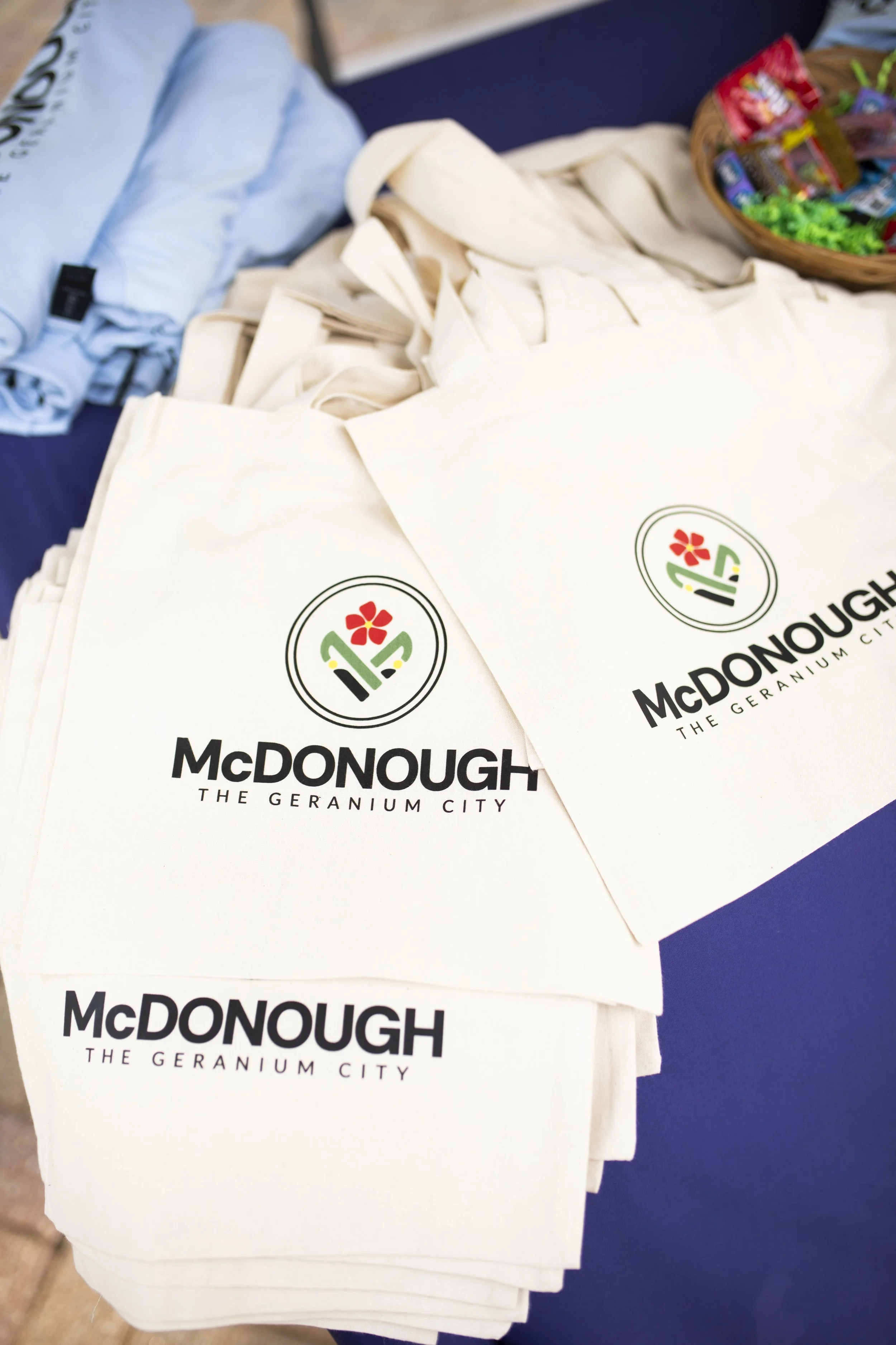 White tote bags with the McDonough logo and text, stacked on a table with some candy in a basket and folded clothing nearby.