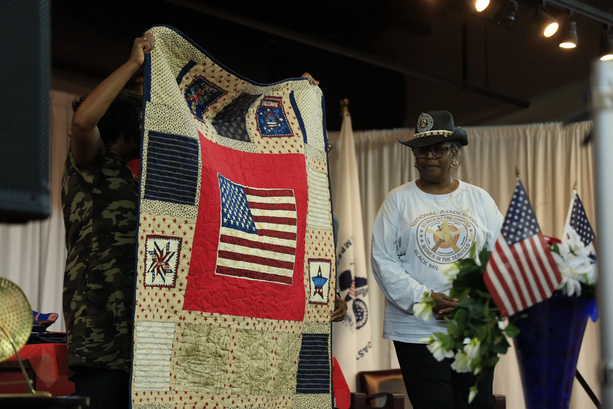 Two women on a stage, one holding a quilt with patriotic patterns and US flags, the other standing beside a stand with American flags and flowers, during a patriotic or commemorative event.