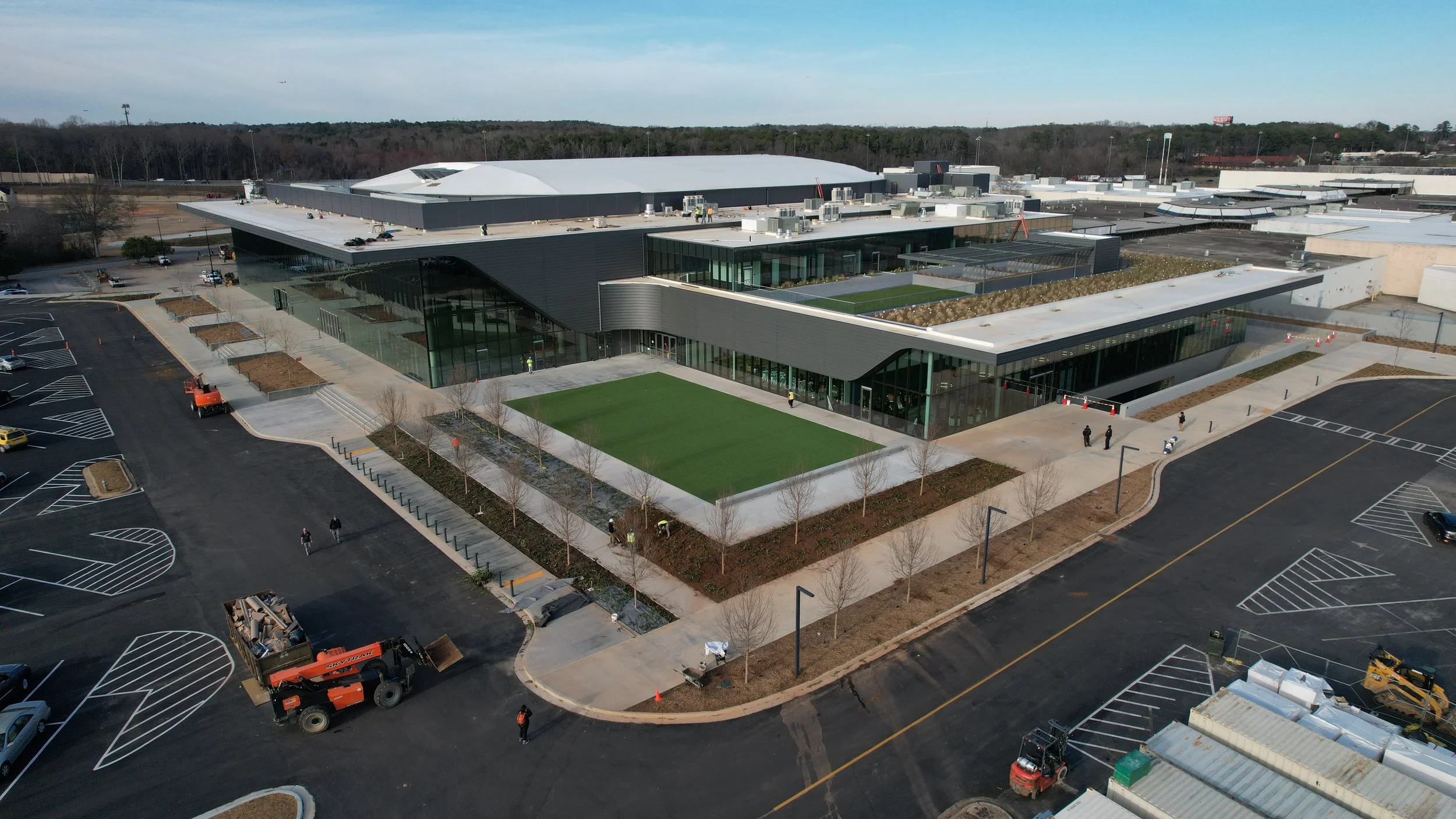 An aerial view of a large modern building with glass walls, surrounded by a parking lot with marked spaces, some trees, and a few people walking nearby. There are construction vehicles and workers present. The building has multiple sections with flat