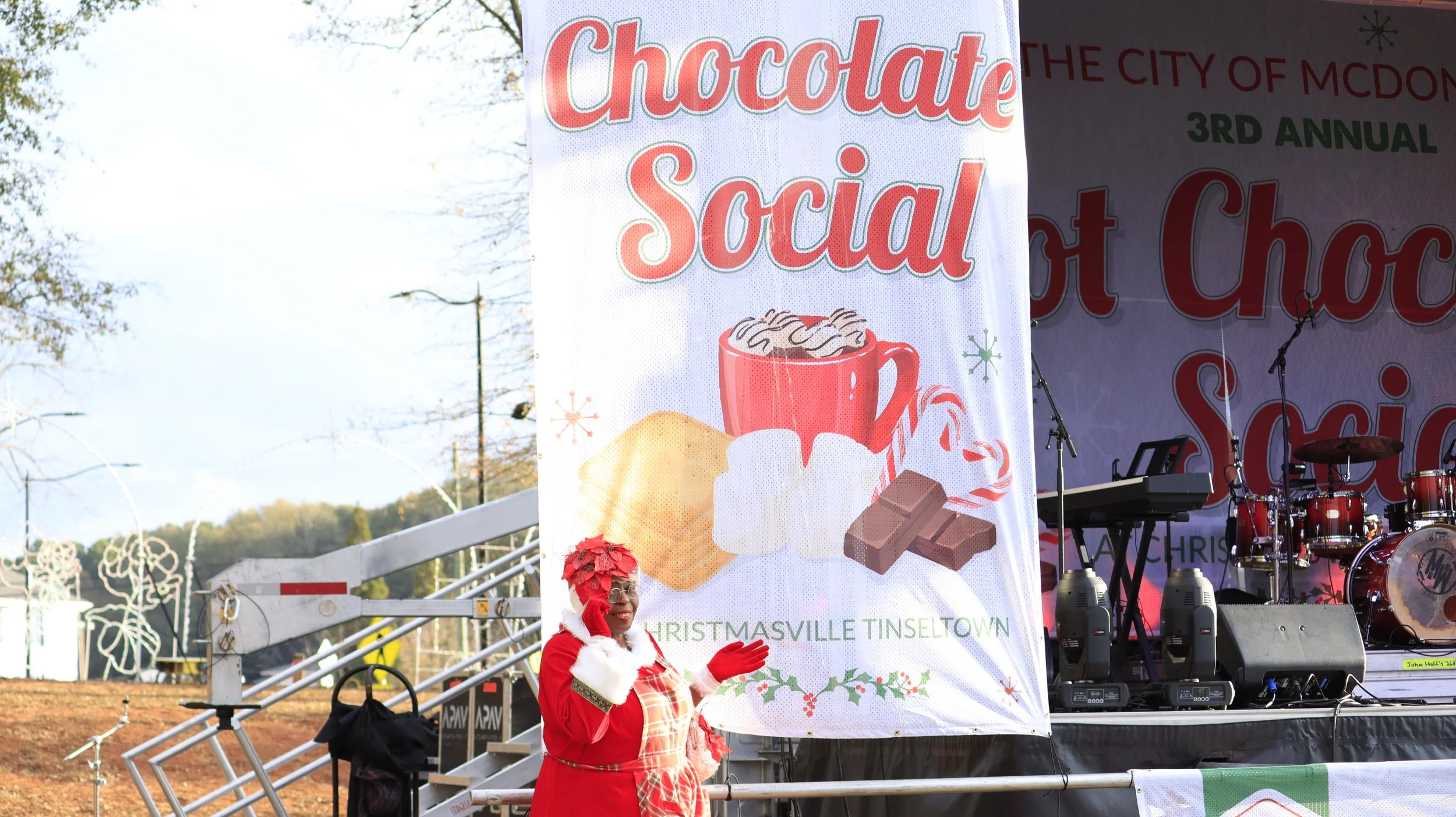 A woman dressed as Mrs. Claus standing next to a large Christmas-themed sign at a holiday event. The sign reads "Chocolate Social" and features images of hot chocolate, marshmallows, a cookie, and chocolates. The background includes a stage with musi