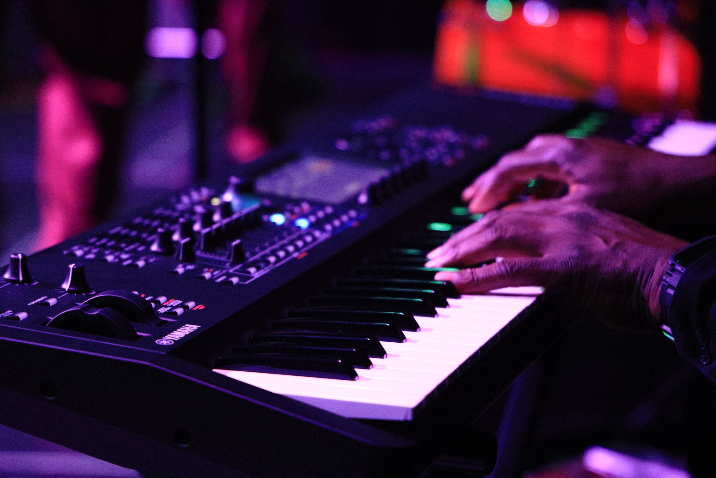 Hands playing a Yamaha keyboard on a dark stage with purple and pink lights.