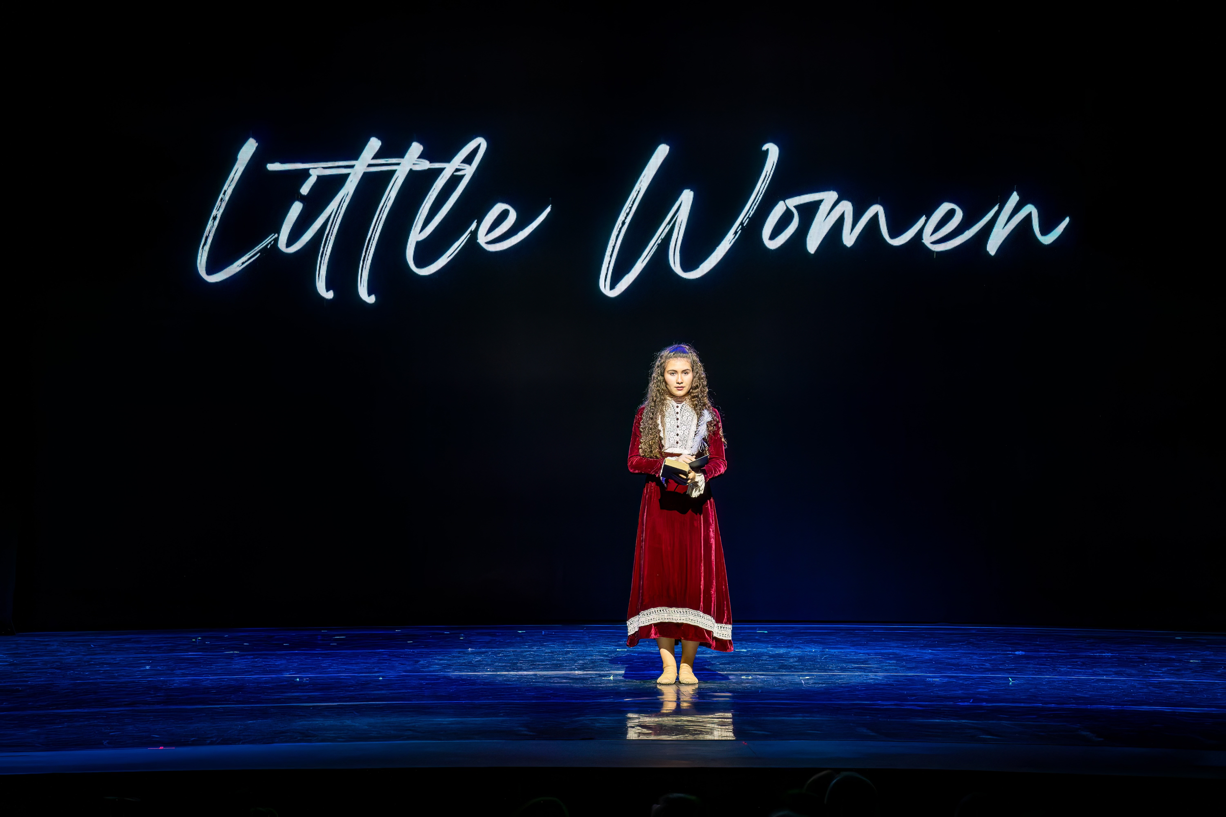 Young girl in a red dress holding books or papers on stage with a backdrop reading 'Little Women' in neon-style font.