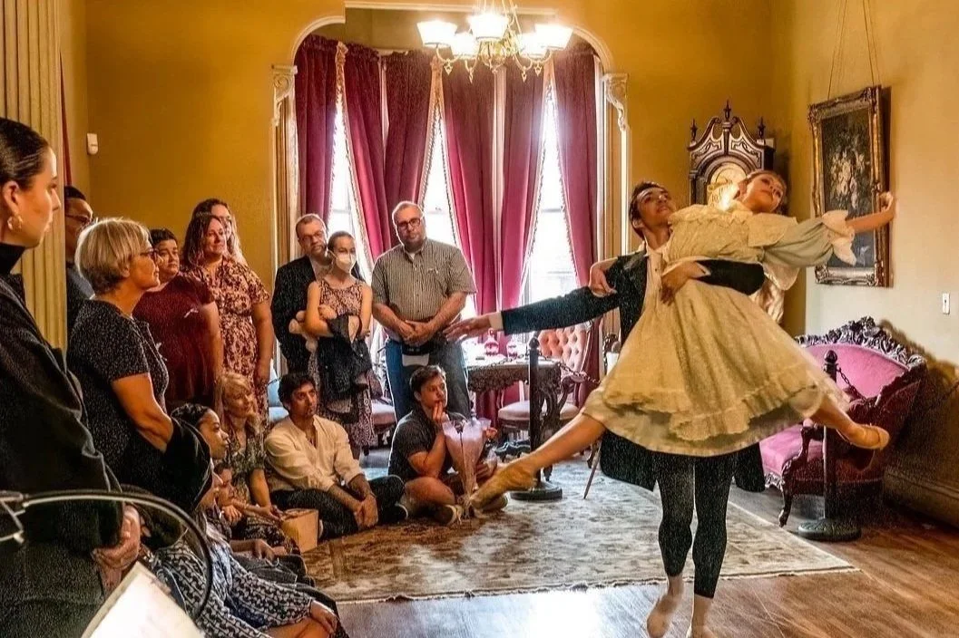 A ballet dancer performing a lift in front of an audience in a living room with vintage decor, velvet curtains, and a chandelier.