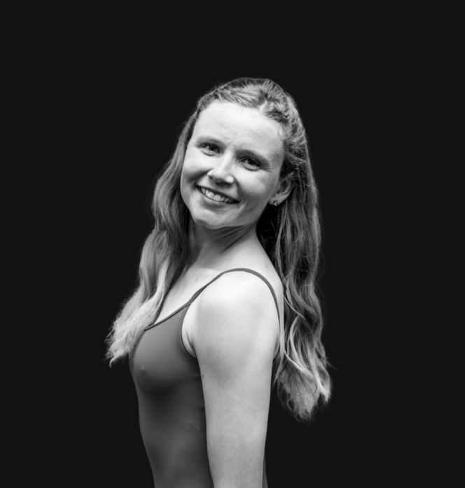 Smiling woman with long blonde hair wearing a colorful dress and a dark shiny jacket, standing indoors against a plain background.