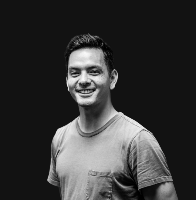 A young man with dark hair smiling, wearing a gray t-shirt, standing indoors in front of a light-colored wall with a mirror and some equipment in the background.