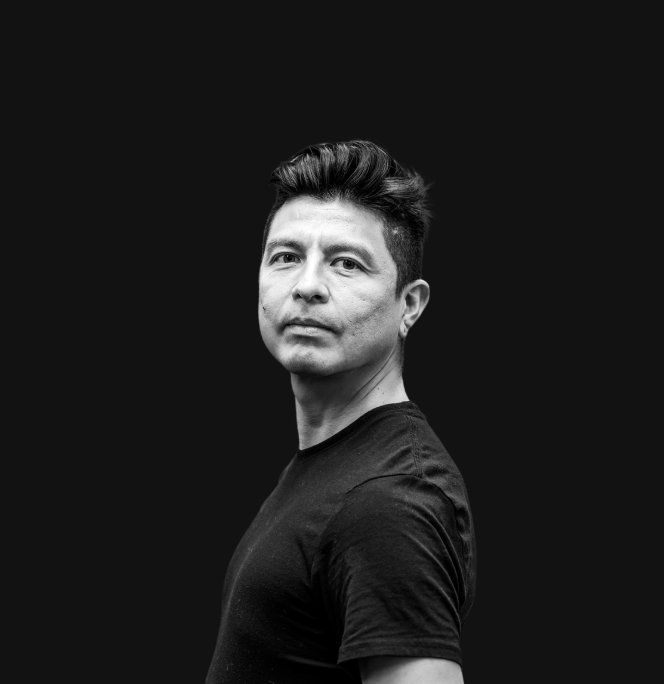 A man with dark hair and light skin wearing a white shirt with a ruffled front and a mustard-colored corduroy jacket, standing against a plain light background.