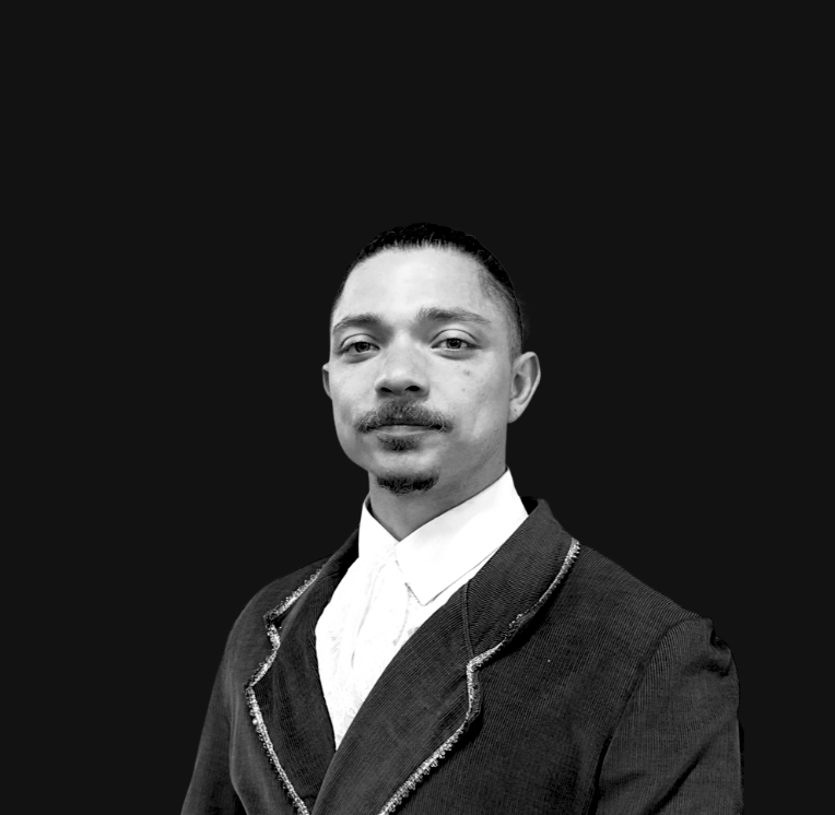 A man with a slicked-back hairstyle, mustache, and goatee wearing a vintage brown suit with a white shirt, standing in front of a mirror in a room with white walls and ceiling-mounted speakers.
