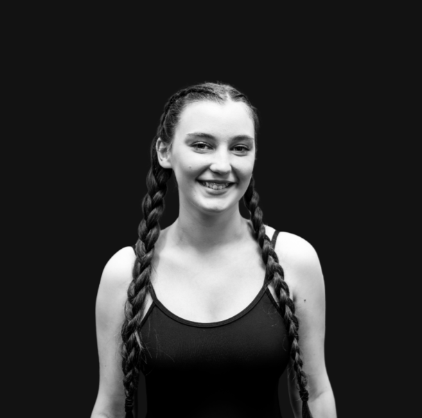 A young girl with long, braided hair, wearing a red dress with black accents, standing indoors and smiling at the camera.