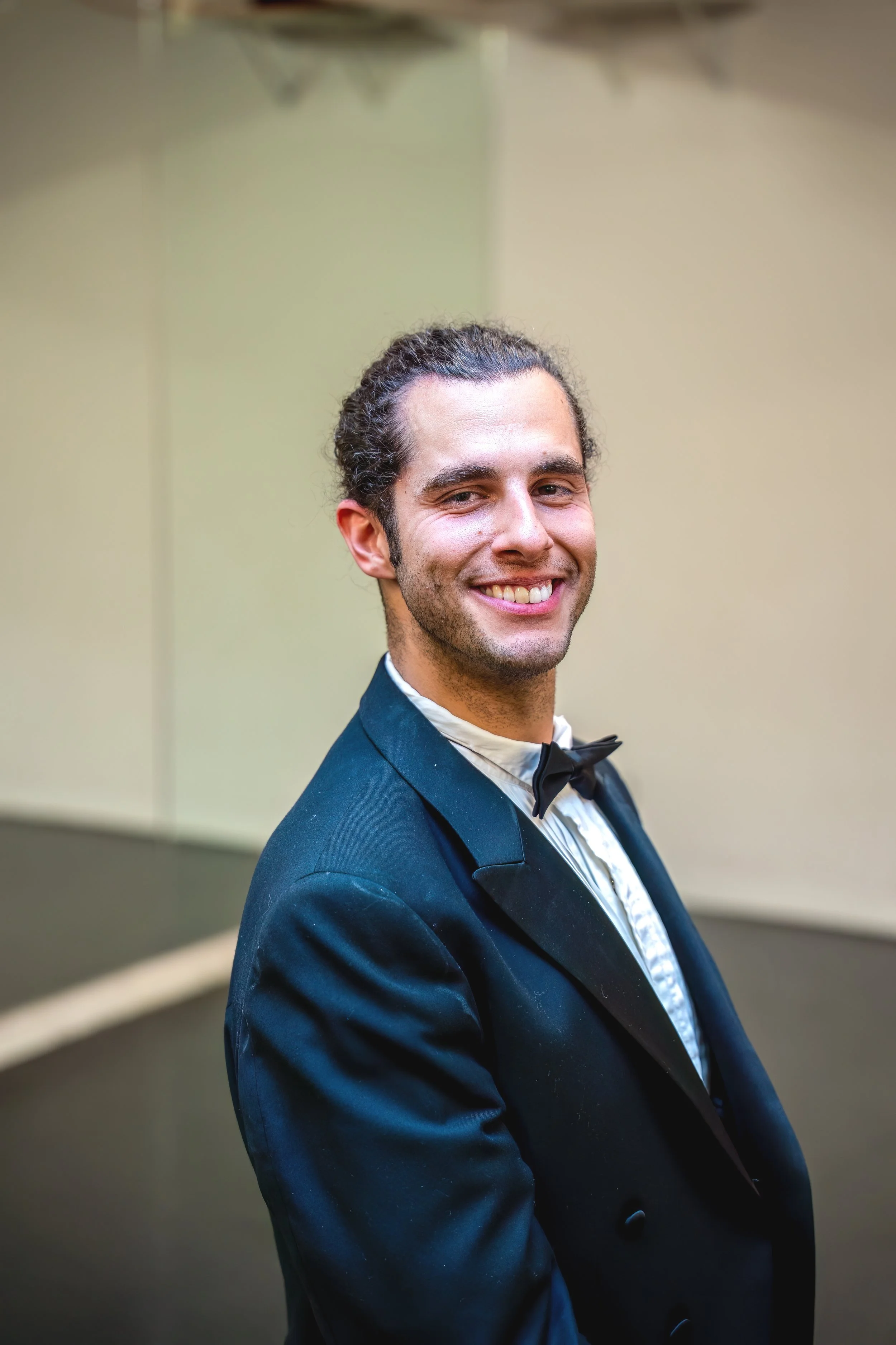 A young man in a tuxedo with a bow tie, smiling and posing in front of a plain background.