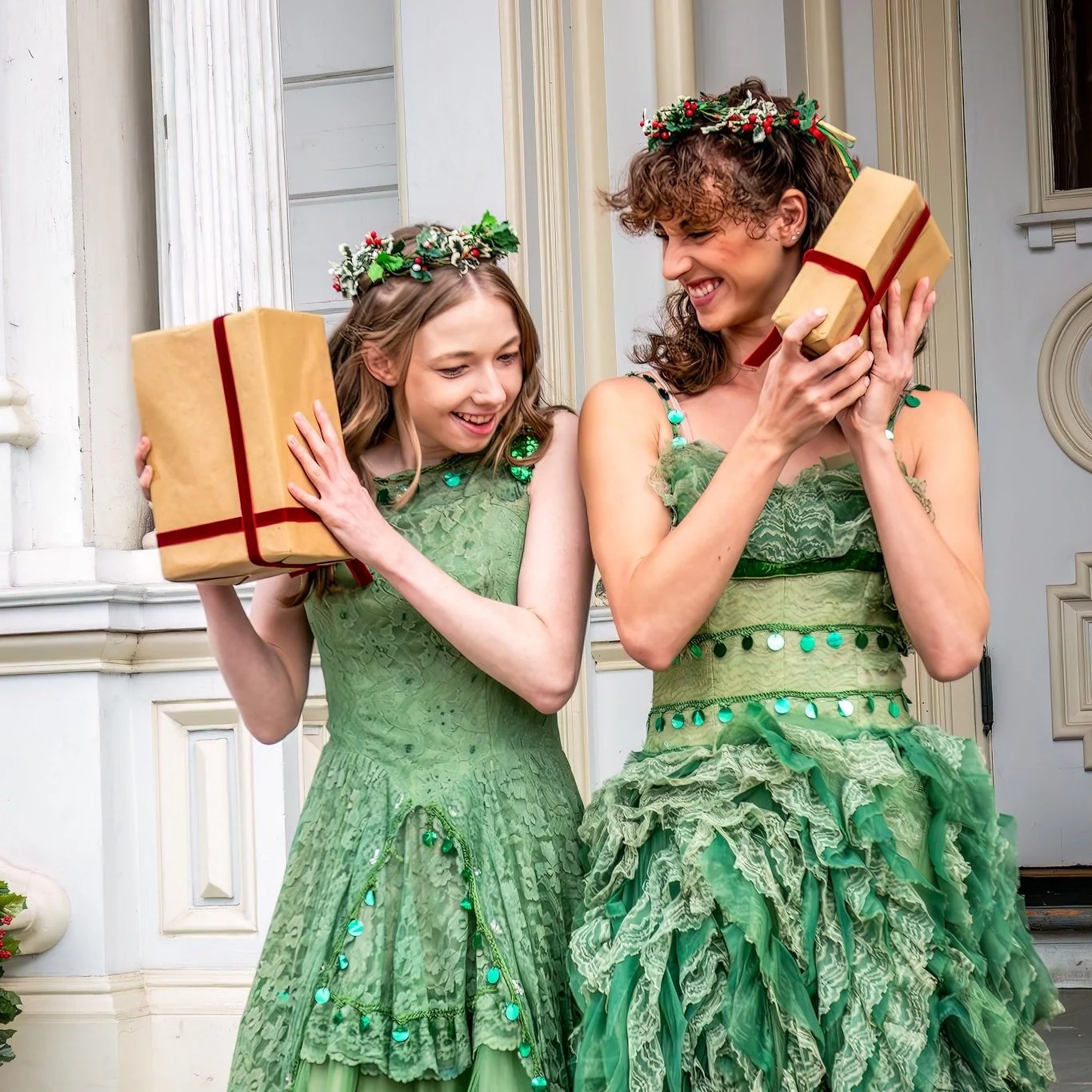 Two women in green dresses and holiday wreath headbands are smiling and holding wrapped gifts, standing in front of a house decorated for Christmas.
