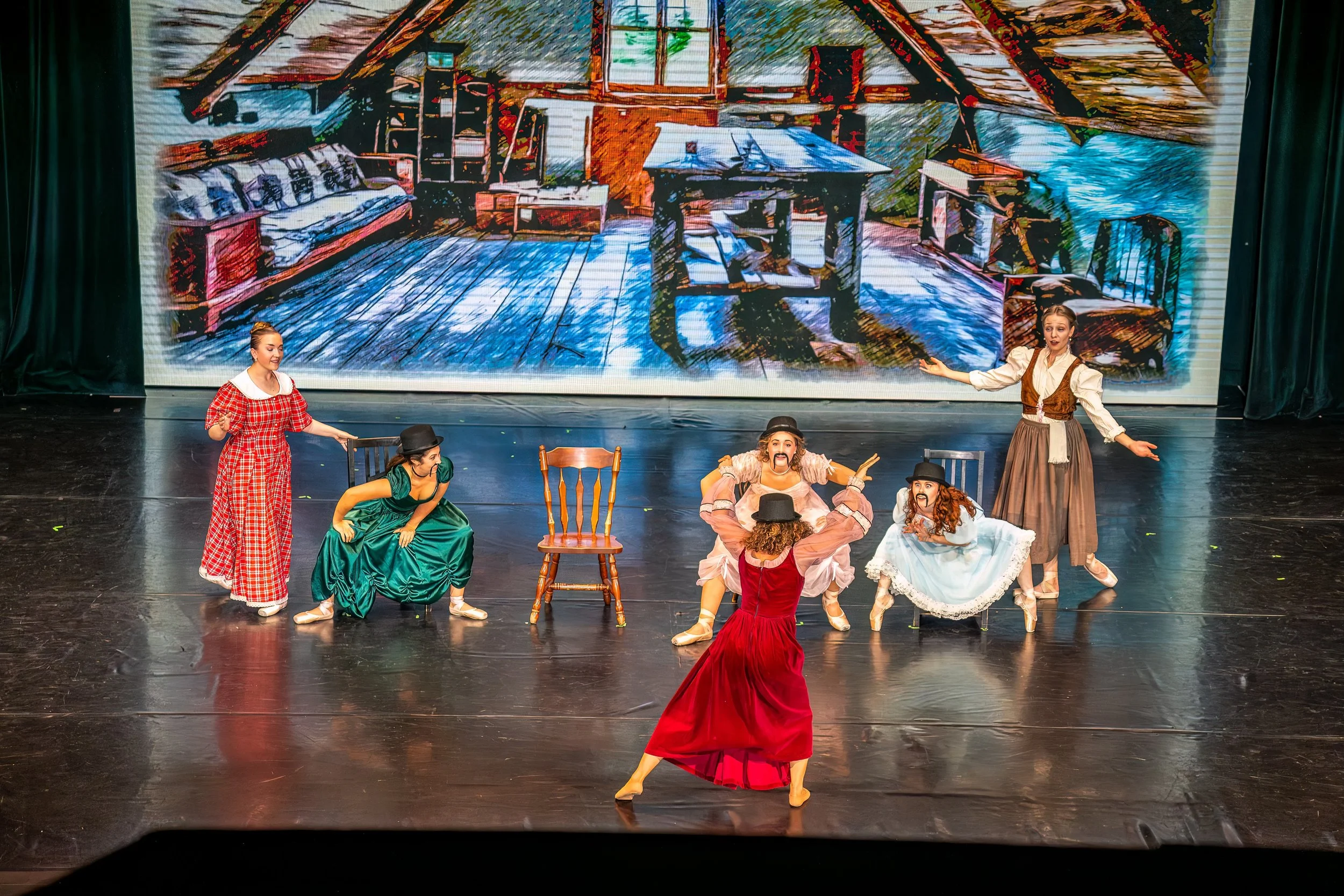 Theater stage with six actors in period costumes performing a play, with a painted backdrop of a rustic interior scene featuring furniture and a view of the outdoors.