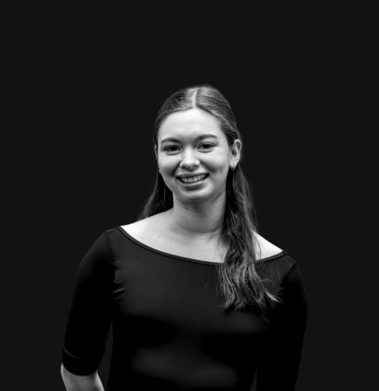 A smiling young woman with long brown hair wearing a black top, standing indoors against a plain background.