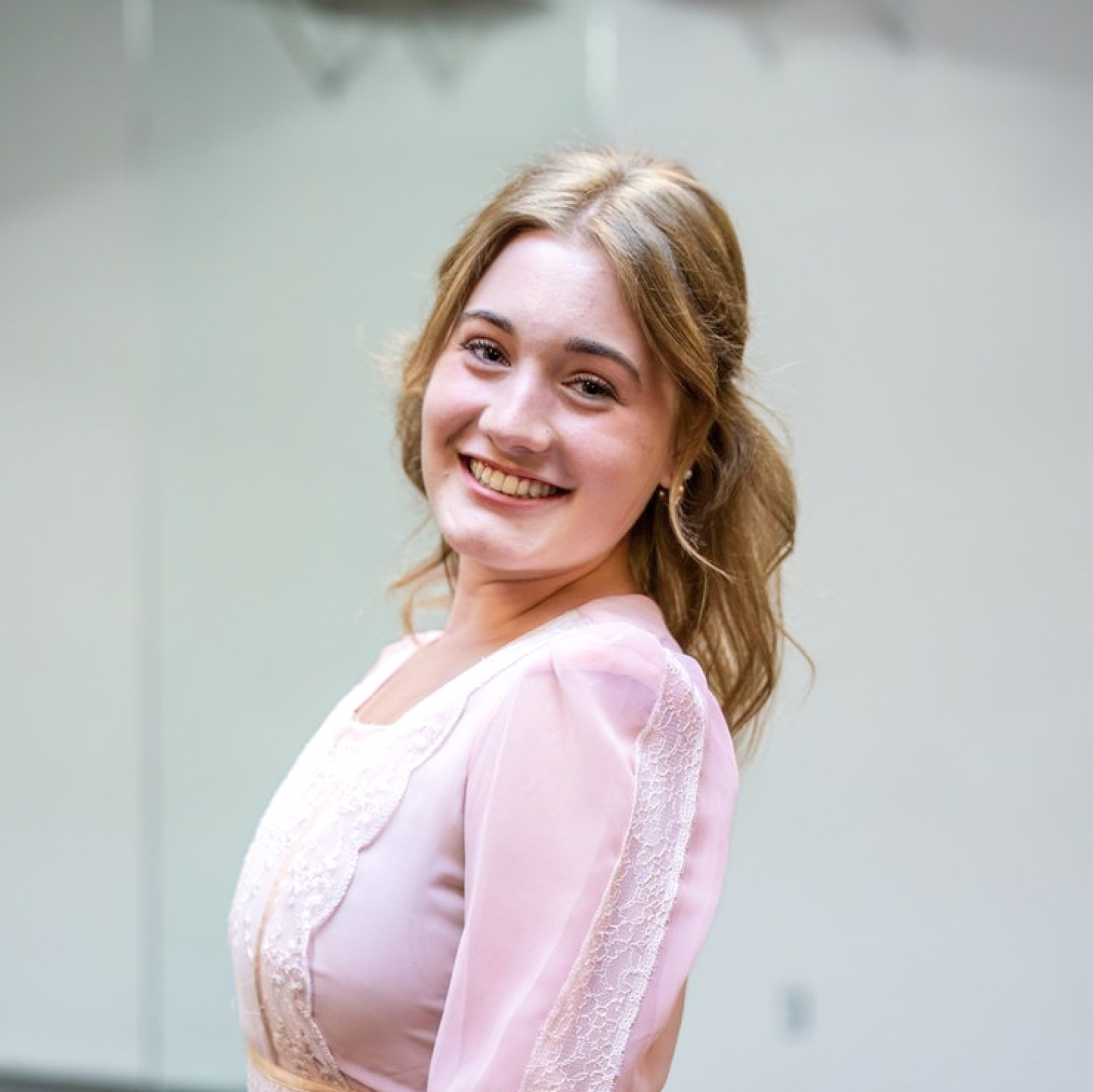 A young woman with light brown hair styled in loose waves, smiling while facing slightly to the right, wearing a pink top with lace details.
