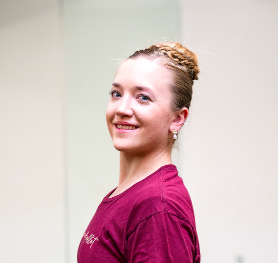 A young woman with light skin and blonde hair styled in a braid, smiling and looking at the camera, wearing a maroon shirt and pearl earrings, against a plain background.