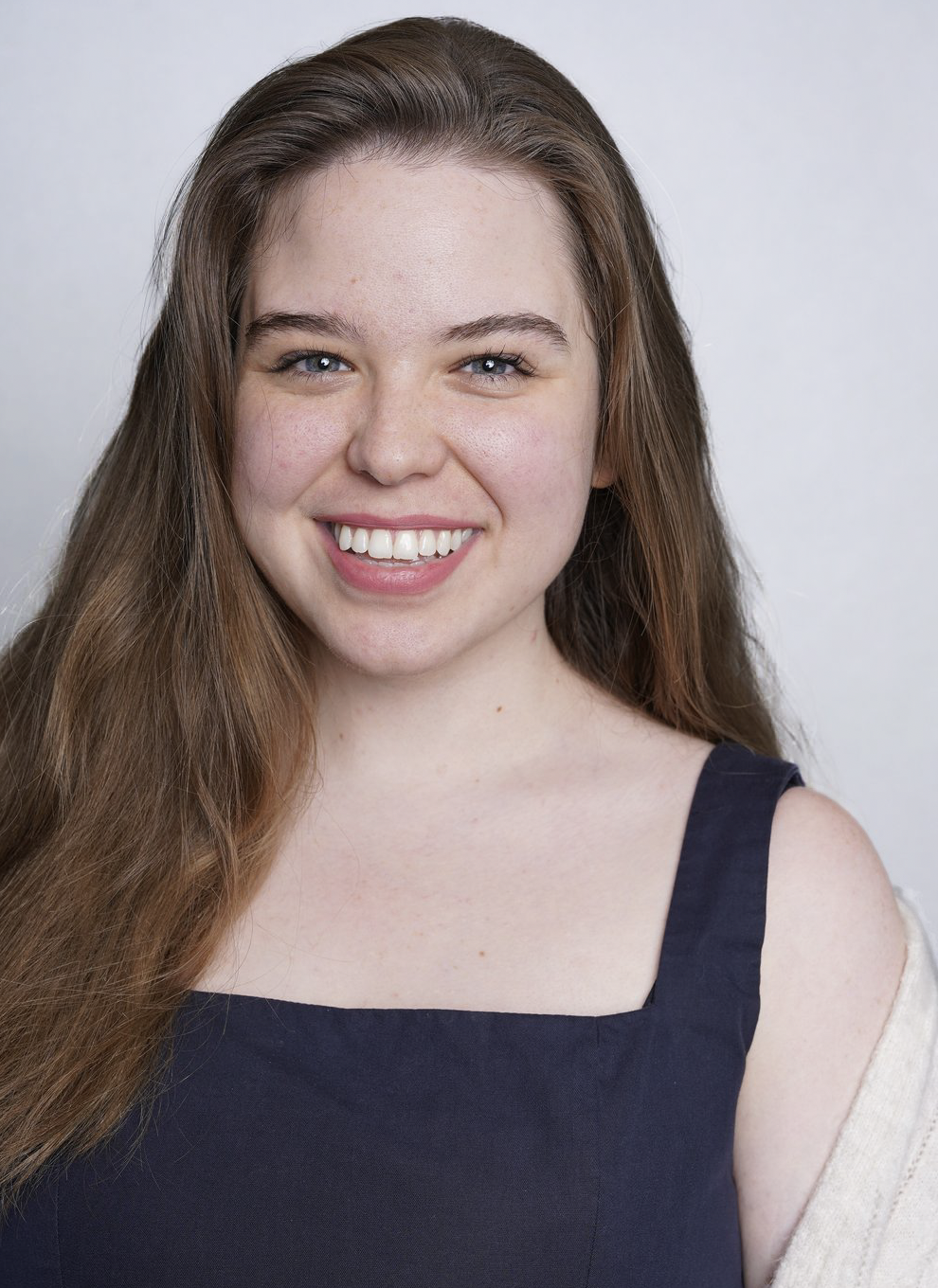 A young woman with long, wavy brown hair, blue eyes, and a bright smile, wearing a black sleeveless top and a light-colored cardigan, standing against a plain white background.