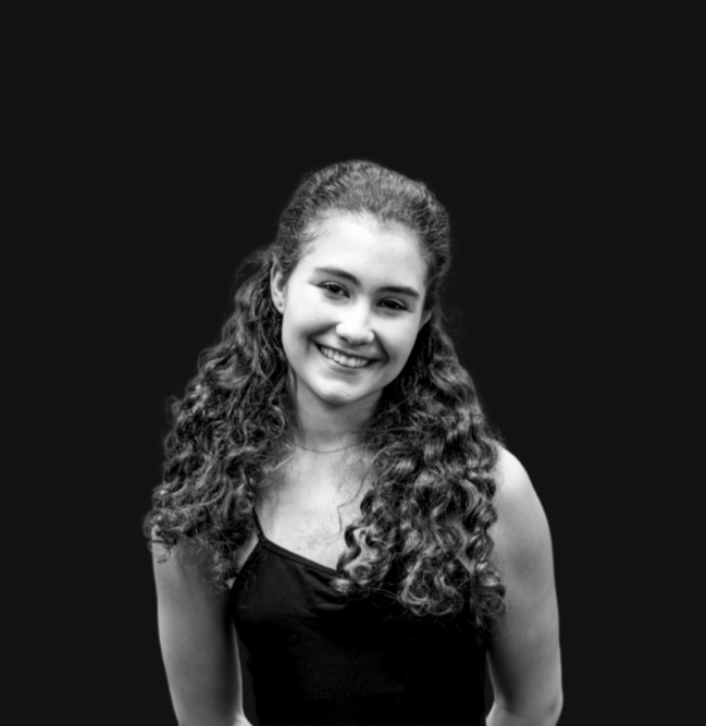 A young woman with curly brown hair, smiling, wearing a black dress with an ornate white lace bib detail, standing against a plain, light-colored wall.