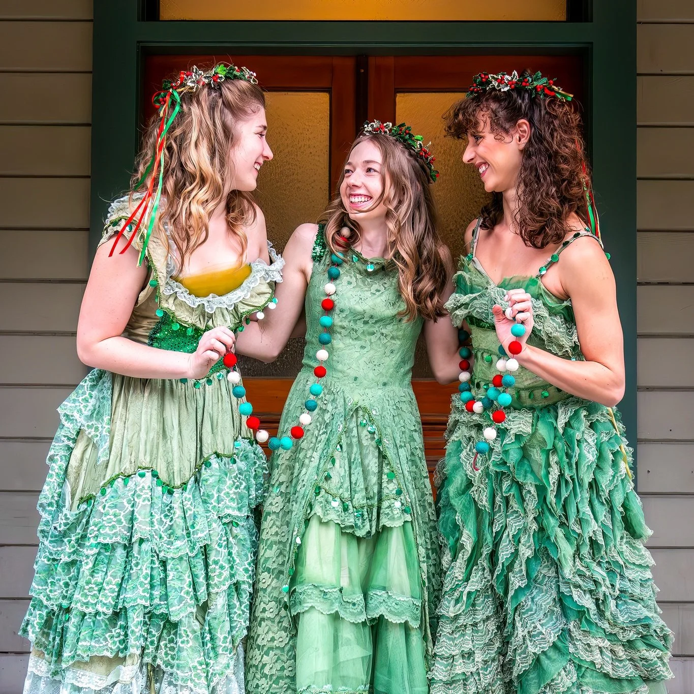 Three women dressed in green fairy tale-style dresses and wearing flower crowns are standing in front of a house door, holding a colorful bead necklace, and smiling at each other.