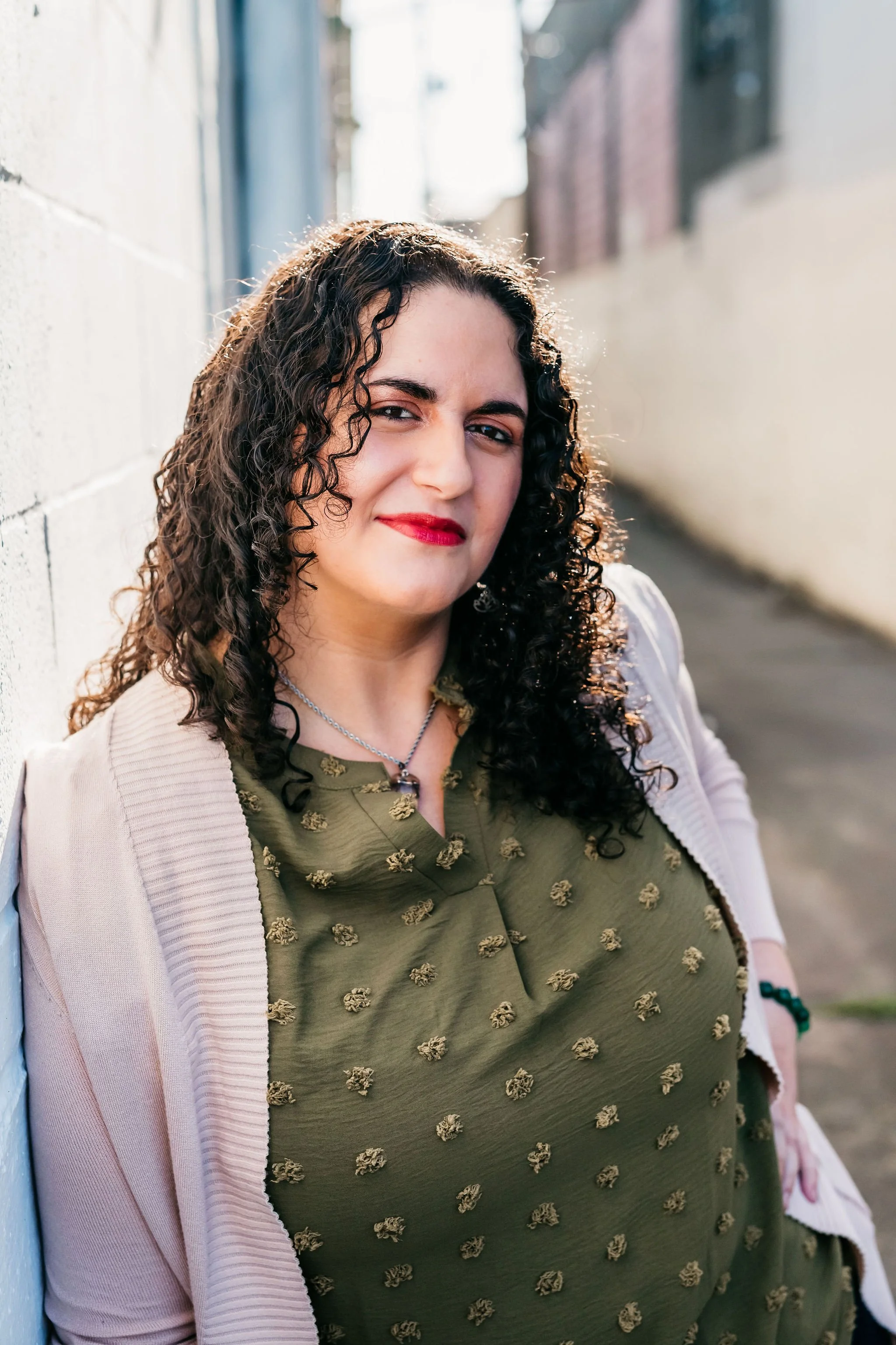 A photo of Jessica looking at the camera and smiling slightly. She has curly brown hair, light skin, and is wearing a green shirt and white cardigan. The background is a blurry building wall.
