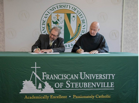 Two men seated at a table signing documents, with a Franciscan University of Steubenville logo and emblem in the background and front, and a green tablecloth with the university's name and slogan.