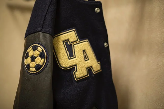 Close-up of a varsity jacket with a football logo patch on the sleeve and large yellow letters 'GA' on the chest.