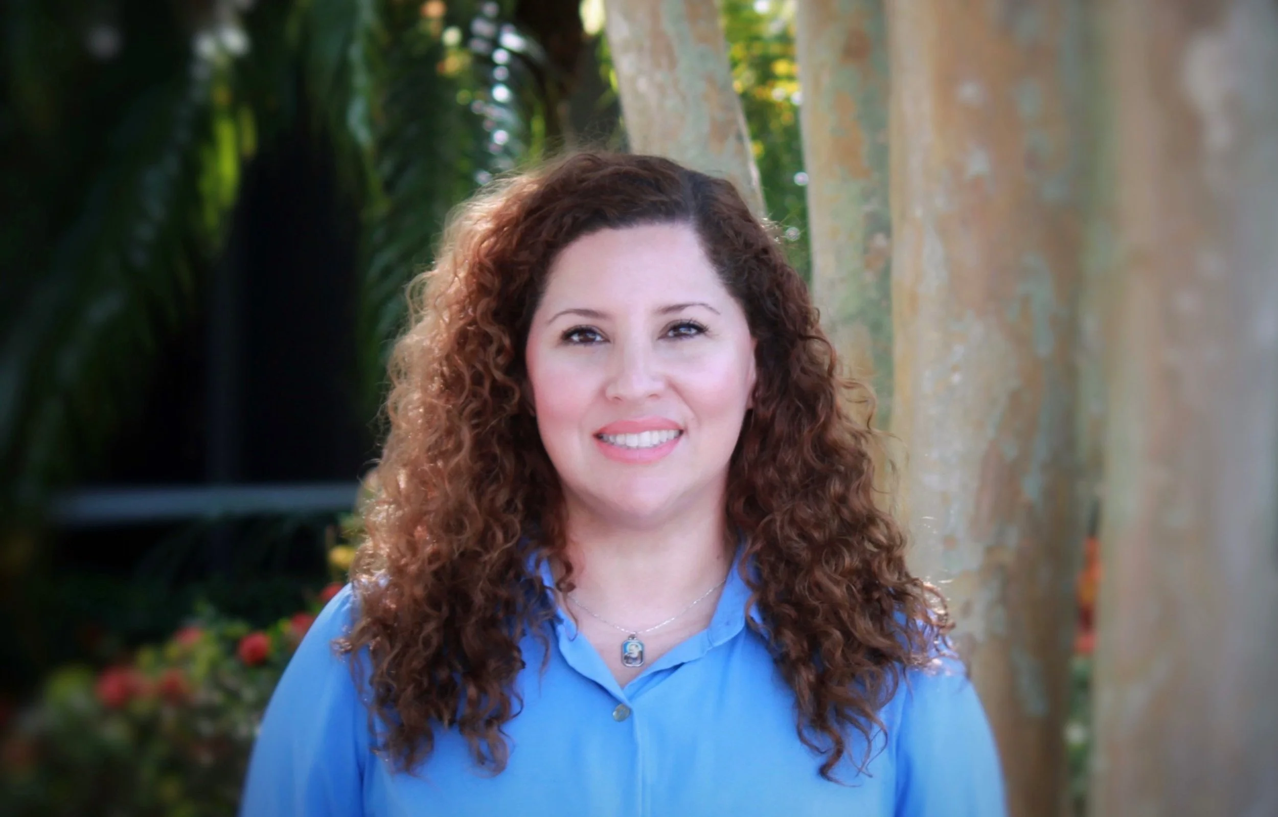 A woman with curly brown hair, wearing a blue shirt and a necklace with a pendant, standing outdoors near trees and greenery.