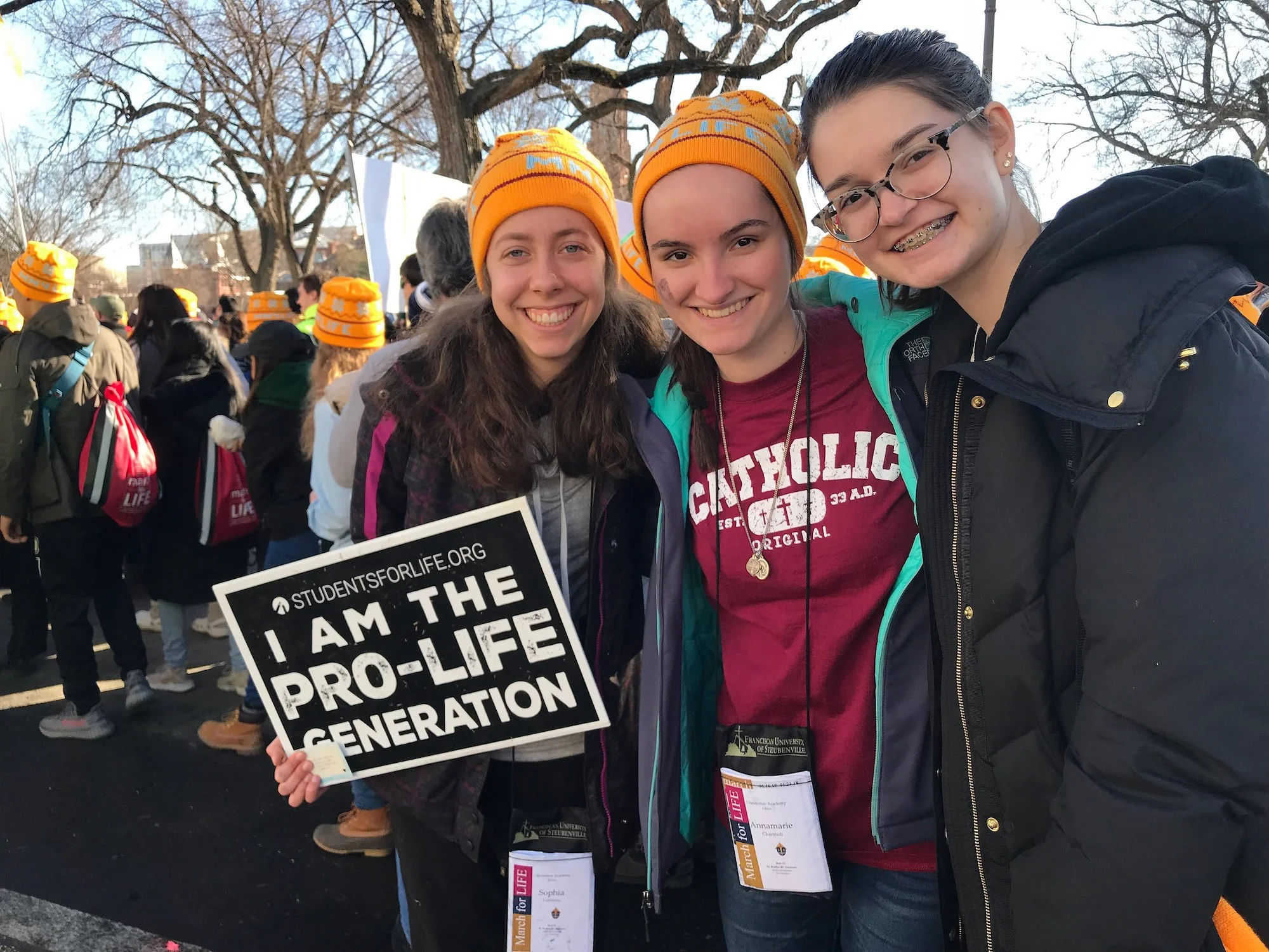 Three young women smiling at an outdoor event, one holding a sign that reads 'I am the pro-life generation', with a crowd and trees in the background.
