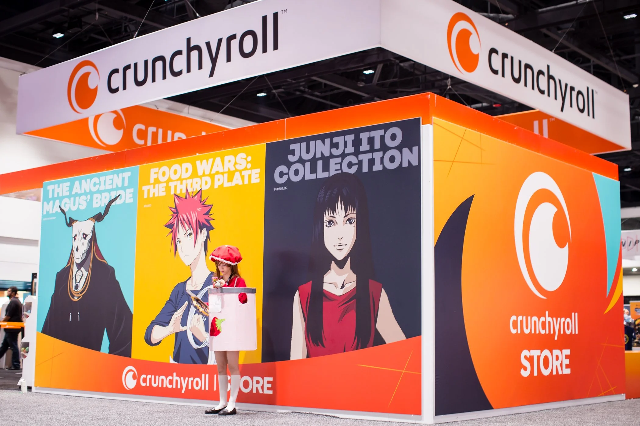 Booth display with large colorful posters of anime characters and the Crunchyroll logo at a convention, featuring a person dressed in a cosplay outfit.