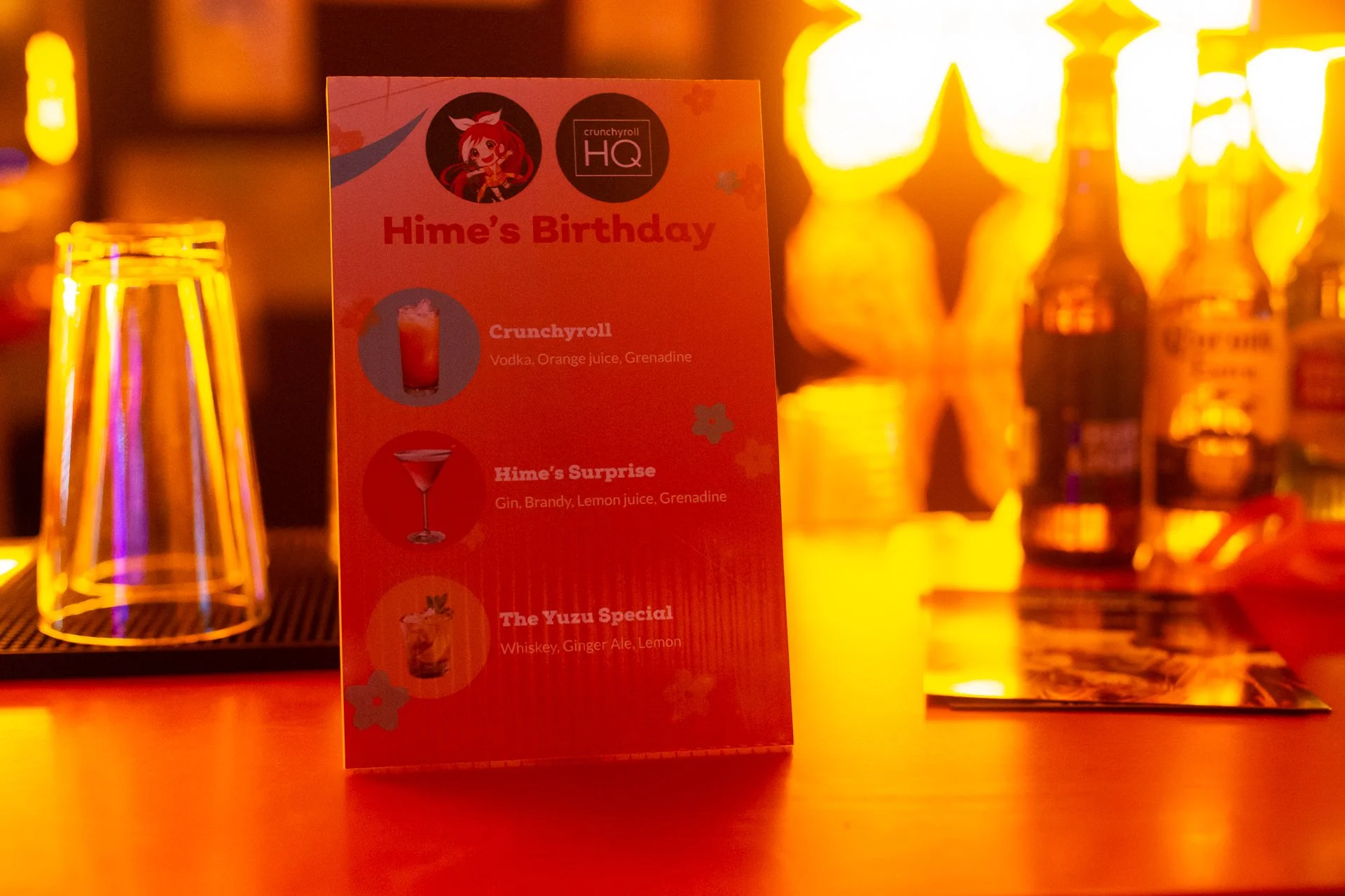 A neon-lit bar counter featuring a birthday drink menu for someone named Hime, with cocktails listed including Crunchyroll, Hime's Surprise, and The Yuzu Special, along with several bottles of beer and transparent drinking glasses.