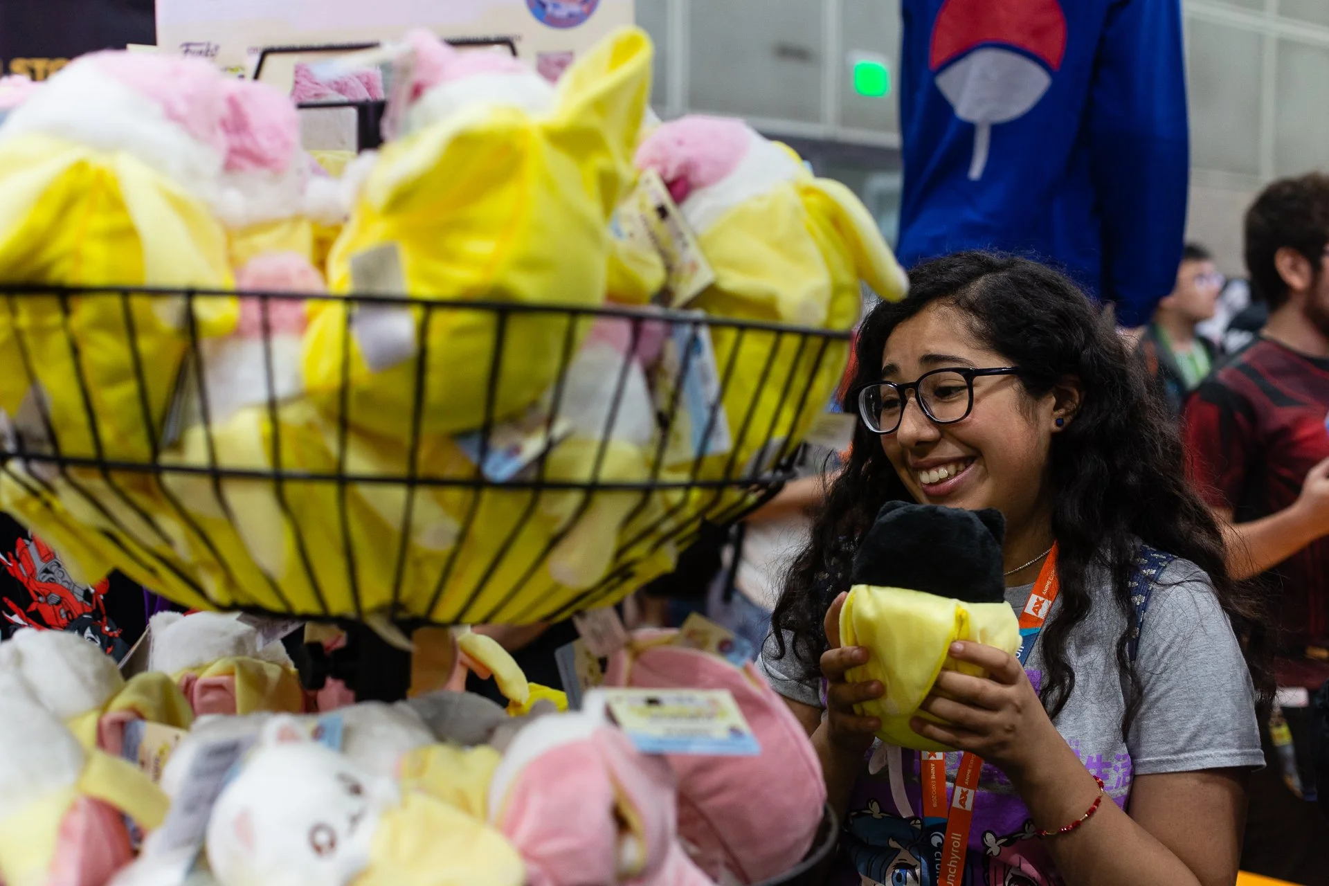 A young woman with glasses smiling and holding a yellow and black plush toy at a store booth filled with pink and yellow plush toys.