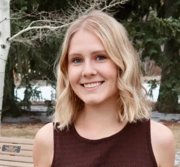 A young woman with shoulder-length blonde hair smiling outdoors near a wooden bench and trees.