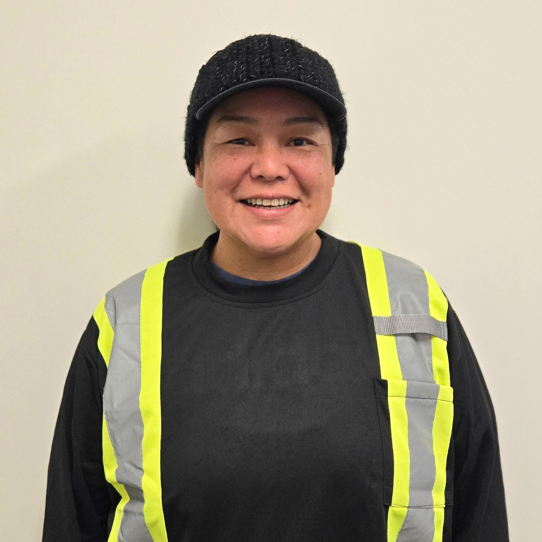 Young woman with long dark hair, green eyes, and light skin, wearing a black hoodie, standing against a wooden wall.
