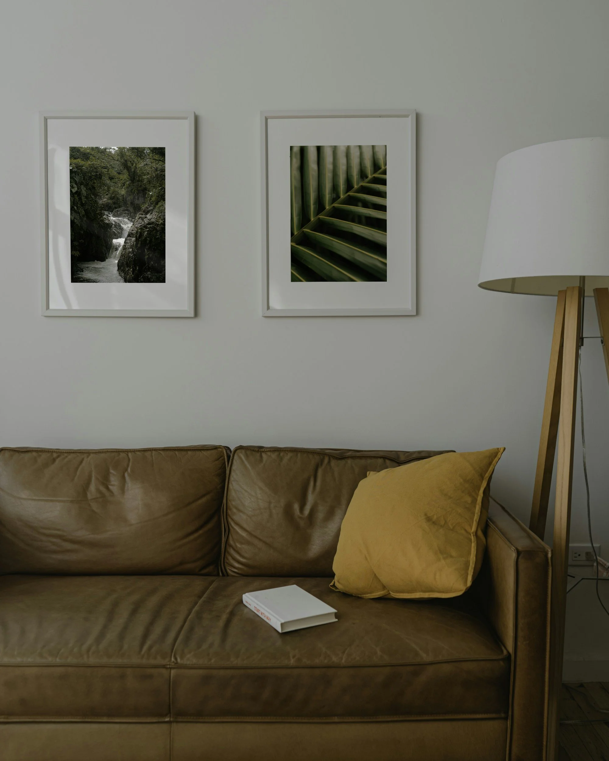Living room with a brown leather sofa, yellow cushion, white framed pictures of nature scenes on the wall, and a white standing lamp.