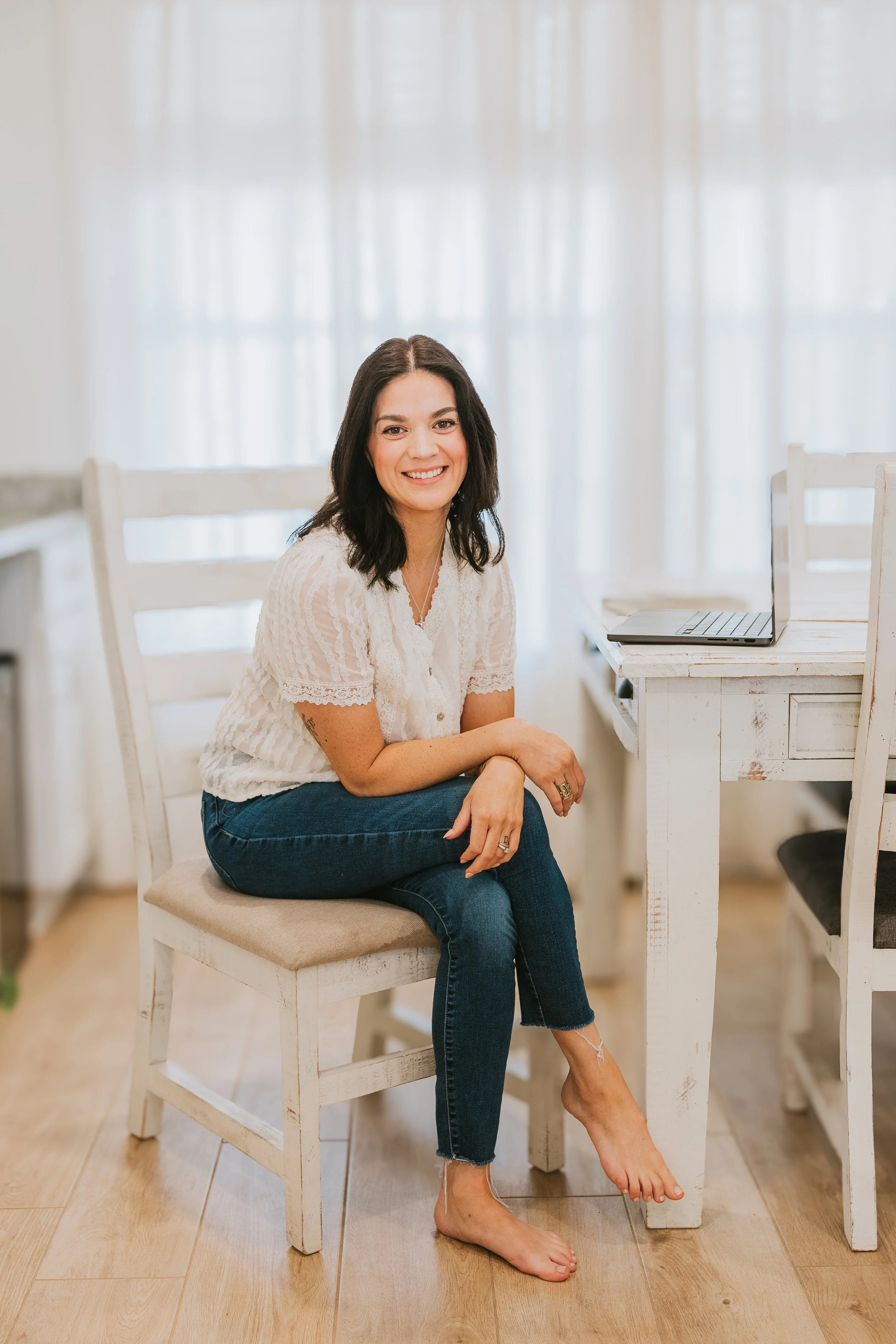 A woman with dark hair smiling, sitting on a white chair at a wooden dining table with a laptop, in a bright room with sheer curtains.