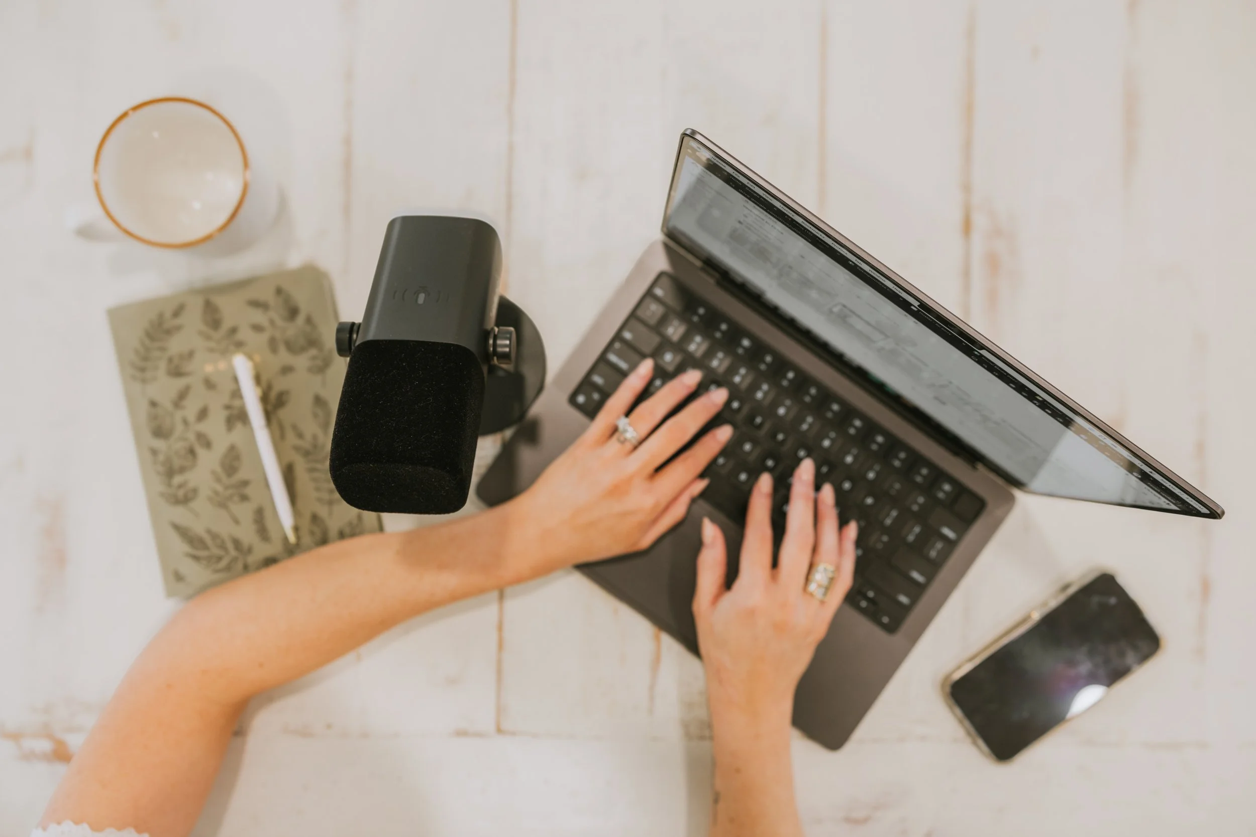 A person's hands typing on a laptop keyboard with a microphone, a smartphone, a notebook, a pen, and a coffee mug on a white wooden table.