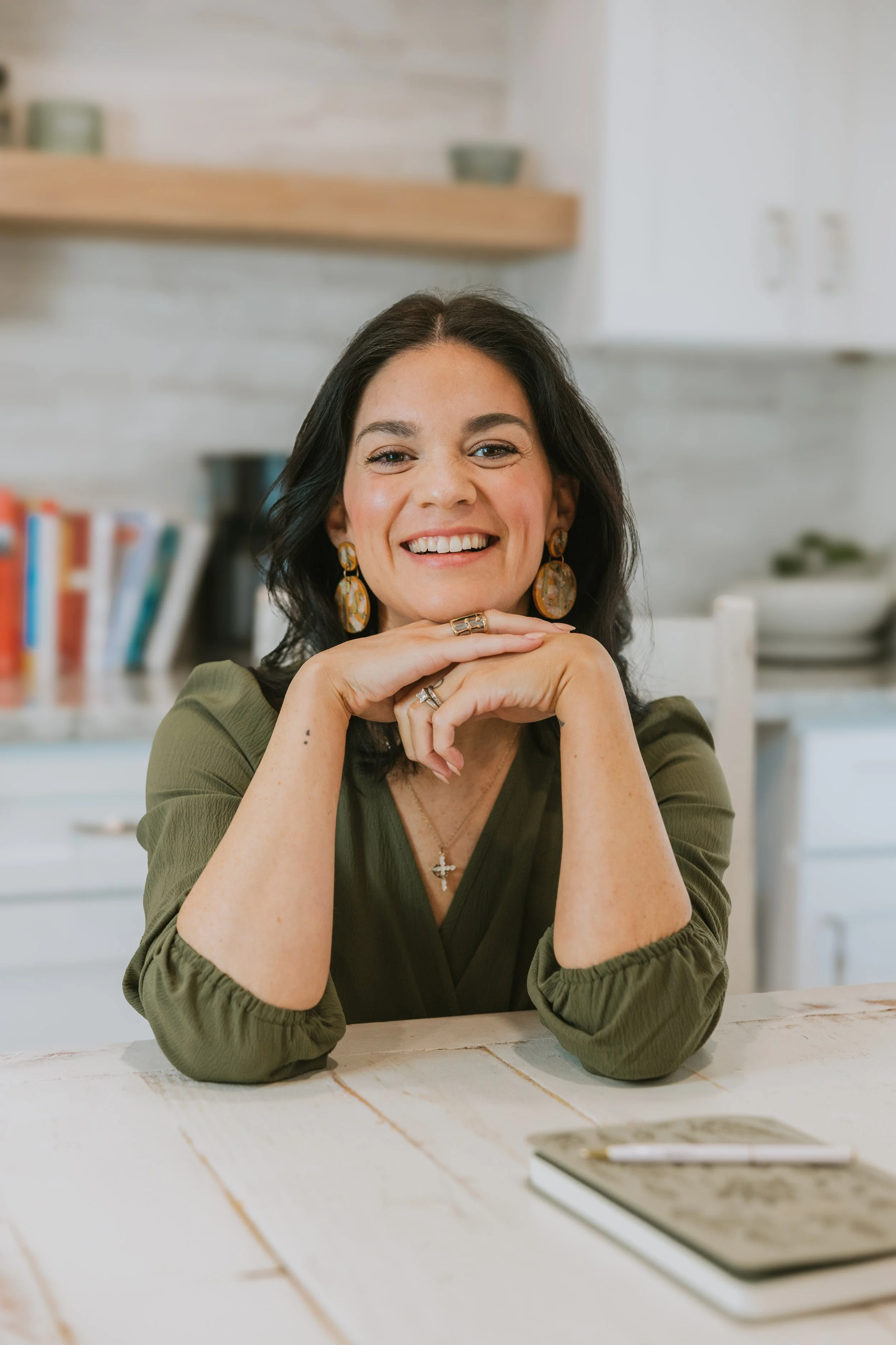 A woman with dark hair and earrings smiling, sitting at a kitchen table with a book and pen in front of her.