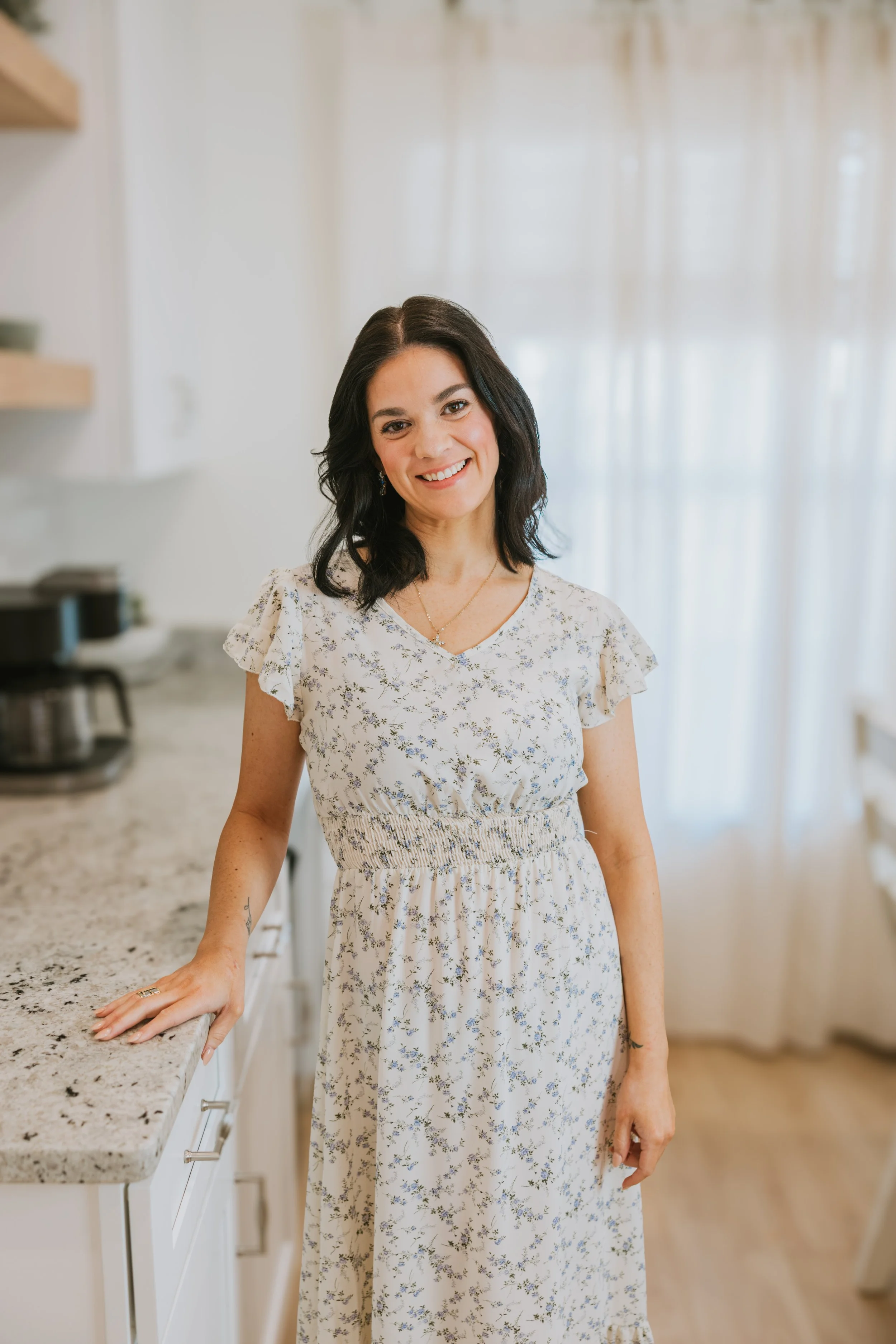 A woman with shoulder-length dark hair in a white floral dress standing in a kitchen with a bright, airy background.