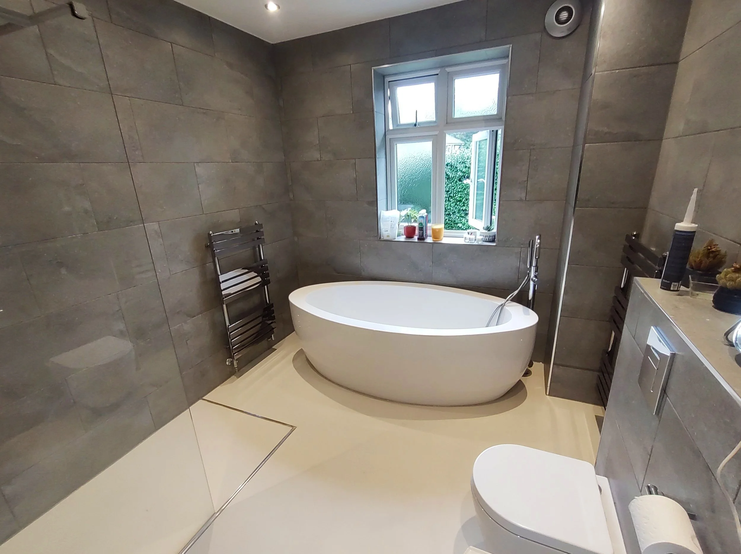 Modern bathroom with a freestanding bathtub near a window, gray tiled walls, a toilet, and a countertop with toiletries.
