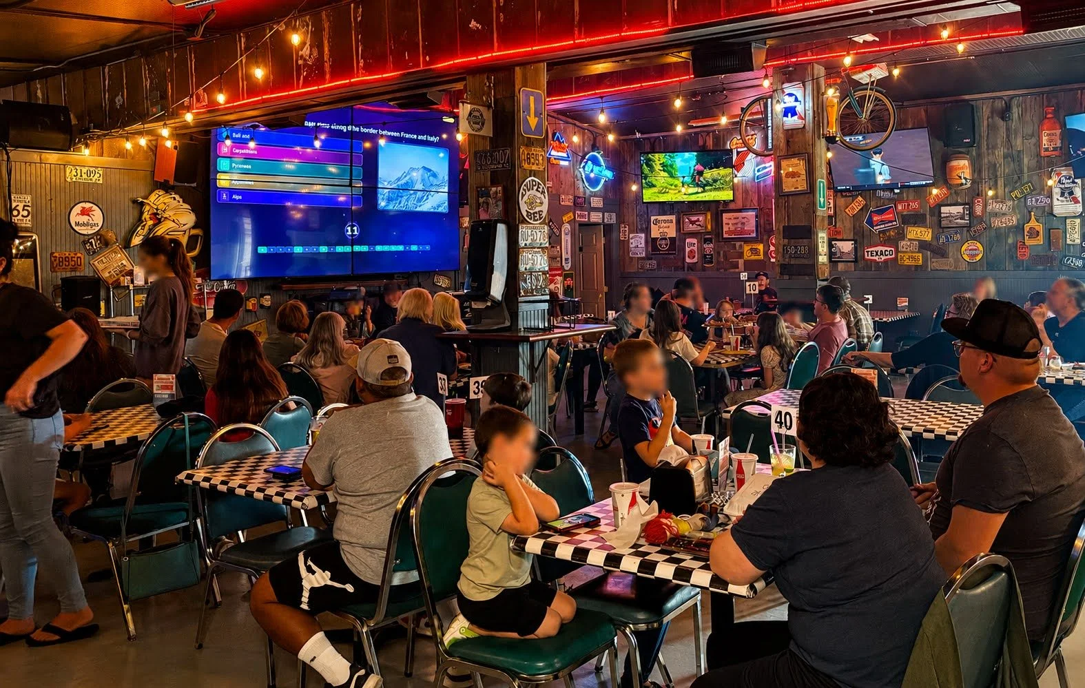 Indoor sports bar with people sitting at tables, watching screens, and a large digital scoreboard. Decor includes vintage signs and neon lights.