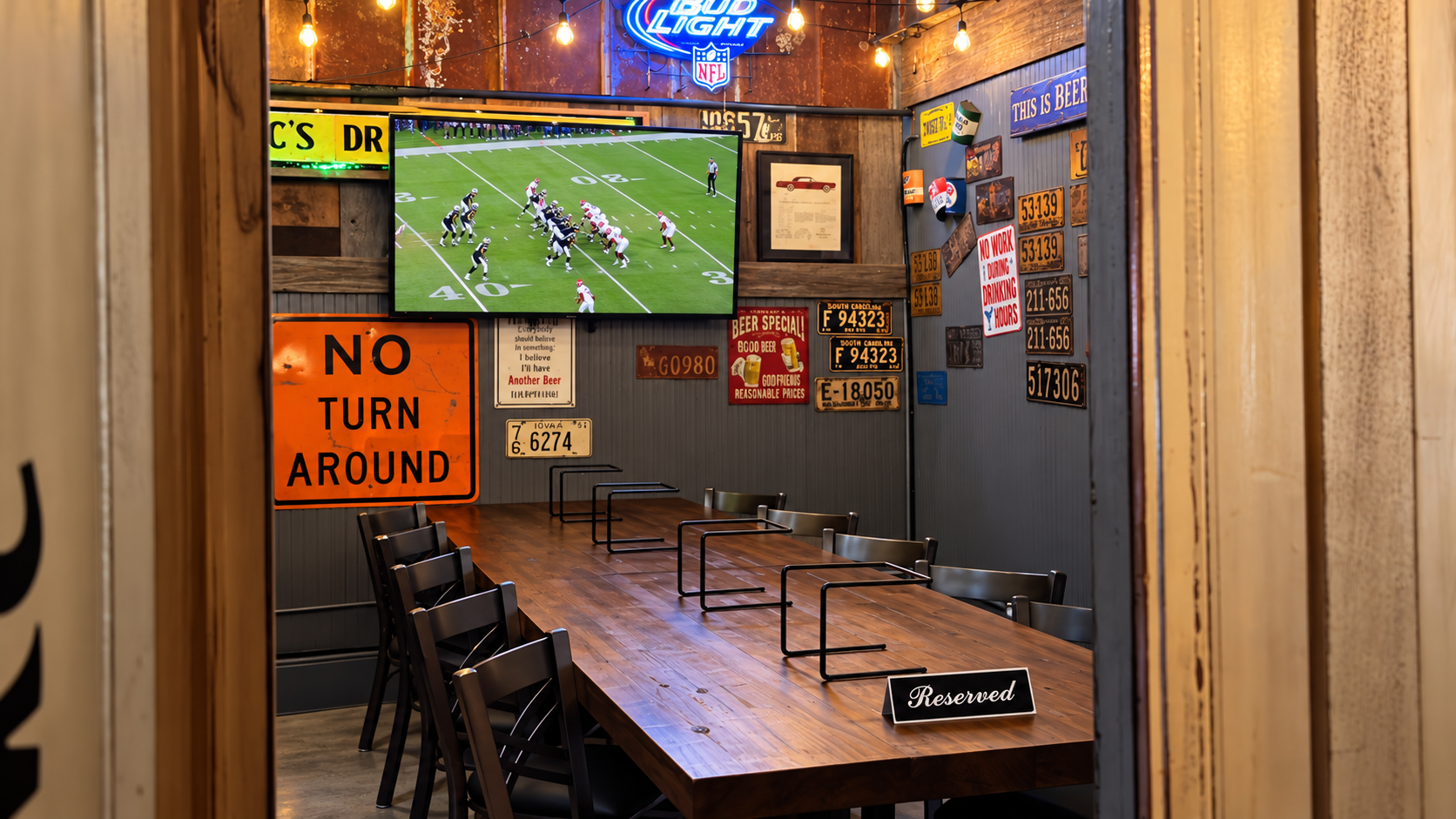 A cozy sports-themed private room with a long wooden table, black chairs, and signs on the wall. A large TV displays a football game, and is surrounded by vintage license plates, signs, and a neon Bud Light sign. The room has warm lighting with string lights hanging from the ceiling.
