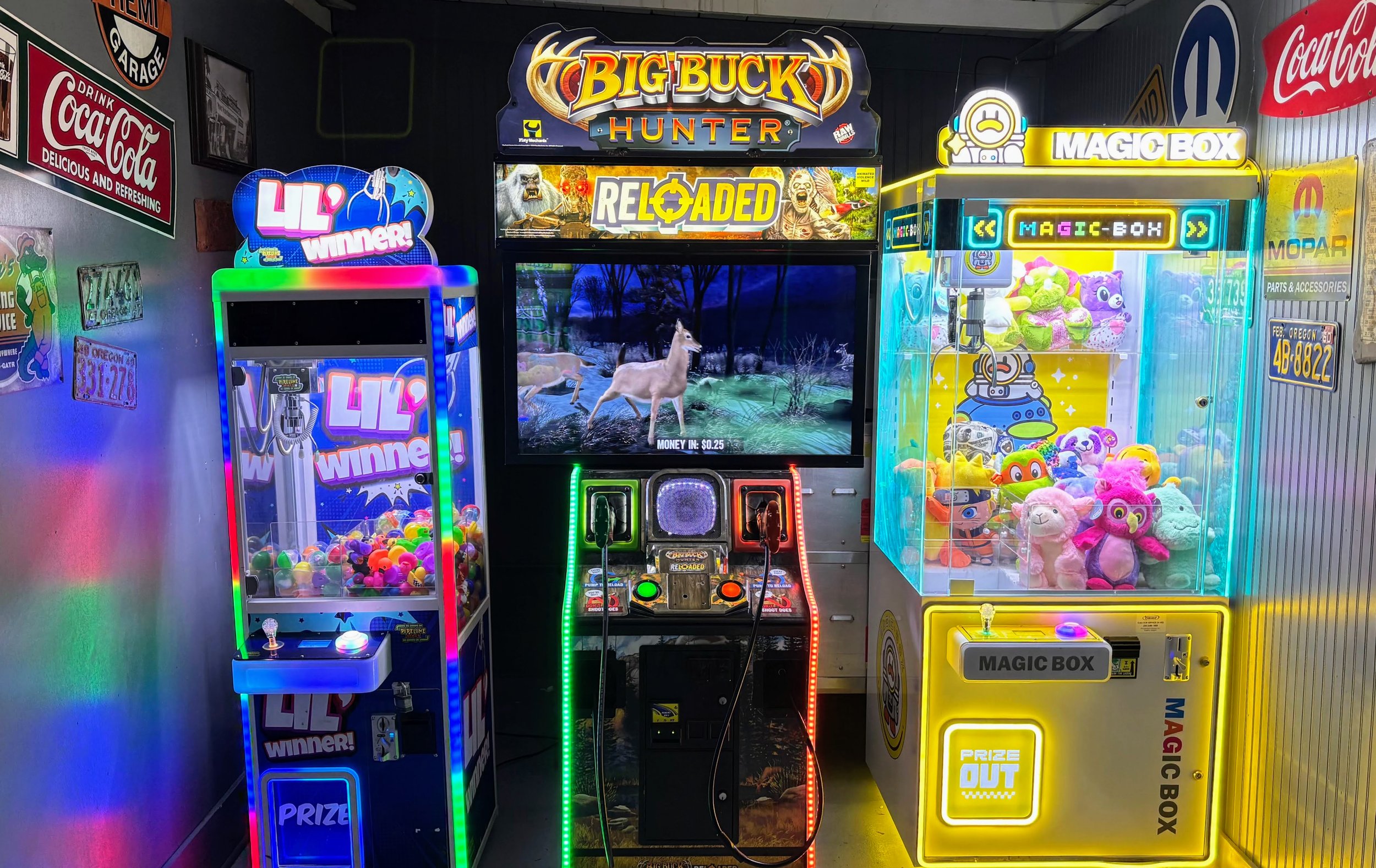 An indoor scene with arcade game machines, including a claw machine filled with plush toys on the right, and a game titled "Big Buck Hunter" in the center. A large screen displays a deer in a natural setting with a "RELOADED" sign above it. There are various neon and illuminated signs and posters on the walls, including a Coca-Cola sign on the left and a "LIL Winner" sign on the left. The scene is colorful and vibrant with a gaming atmosphere.