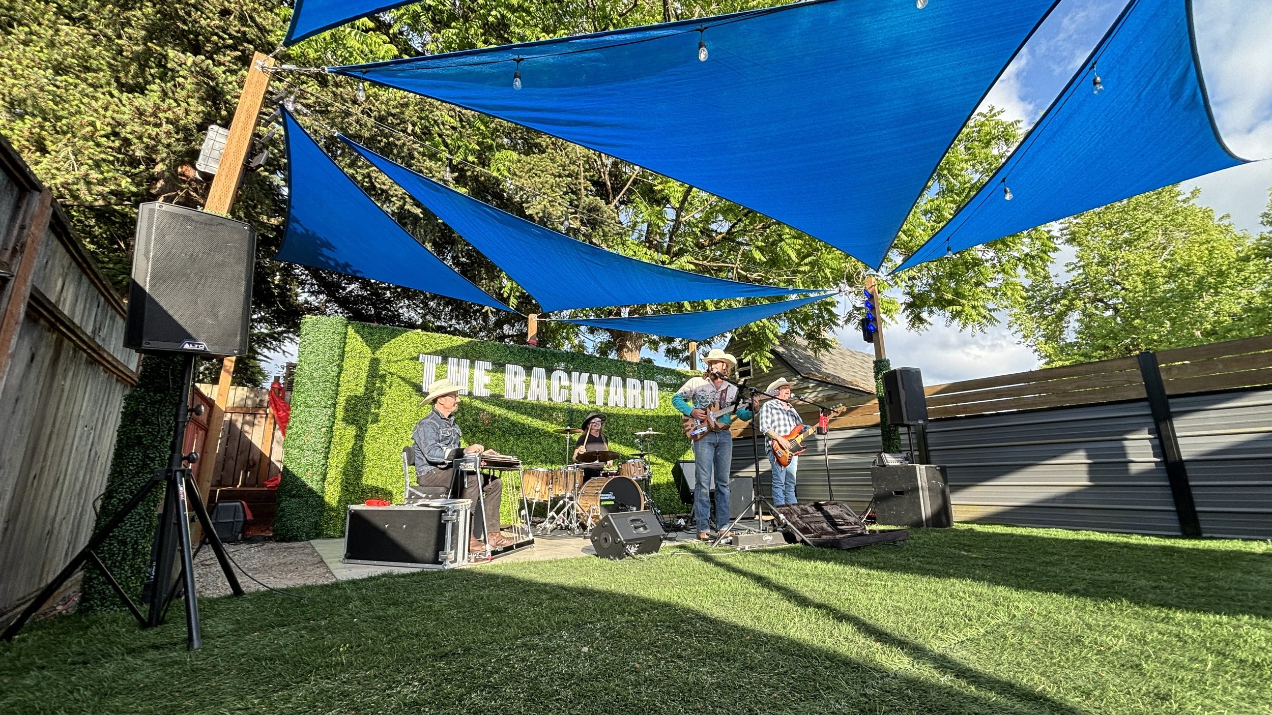 A band of four musicians performing outdoors under blue shade sails, with a green hedge backdrop reading 'THE BACKYARD' in white letters. The setting is in a backyard with trees and a wooden fence, featuring audio equipment and speakers.