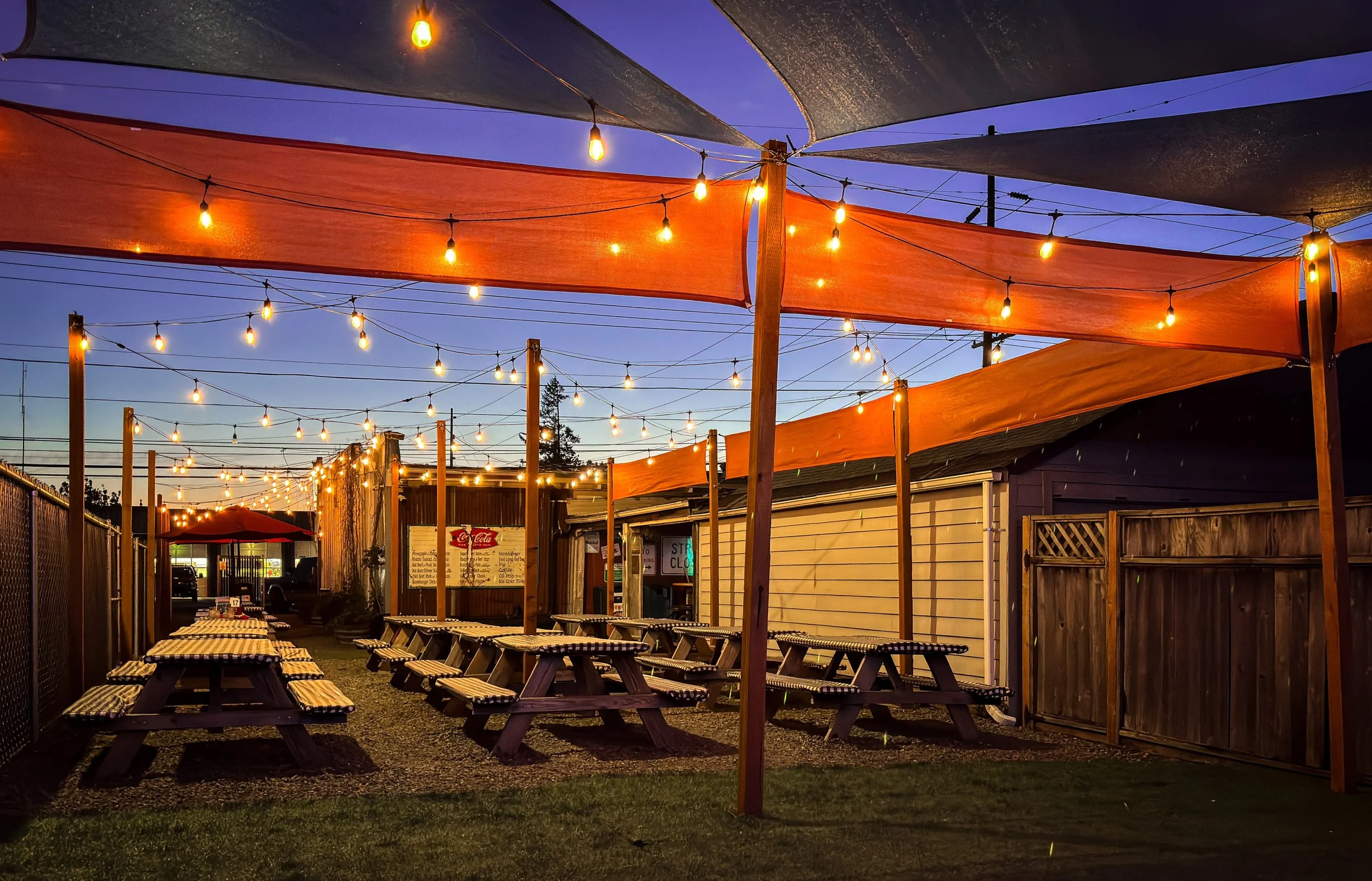 Outdoor dining area at dusk with string lights, picnic tables with checkered tablecloths, and colorful fabric sunshades overhead.