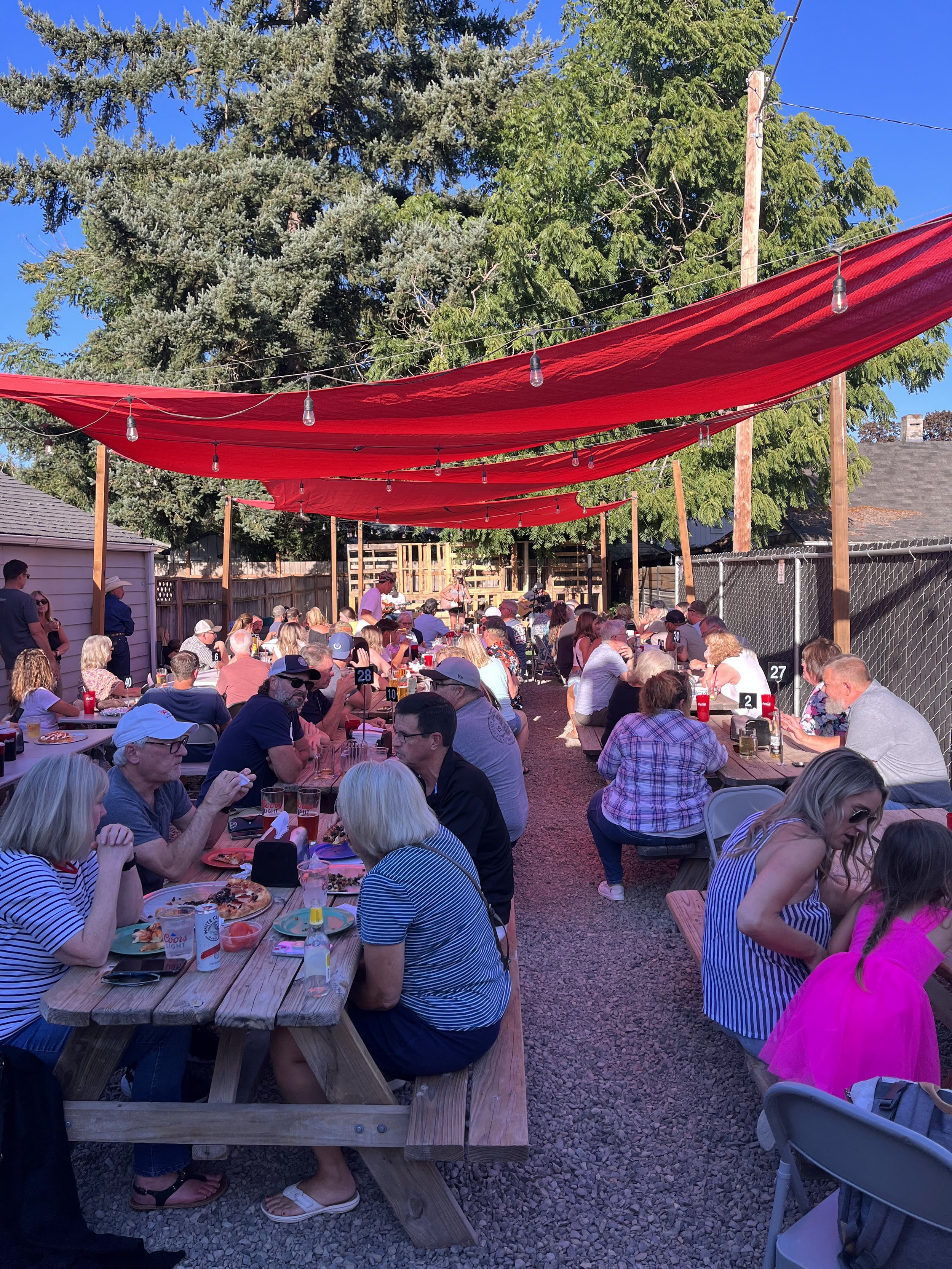 A large outdoor gathering of people seated at long picnic tables under red shade canopies, with food and drinks on the tables, and a band performing on a stage in the background, surrounded by trees and a clear blue sky.