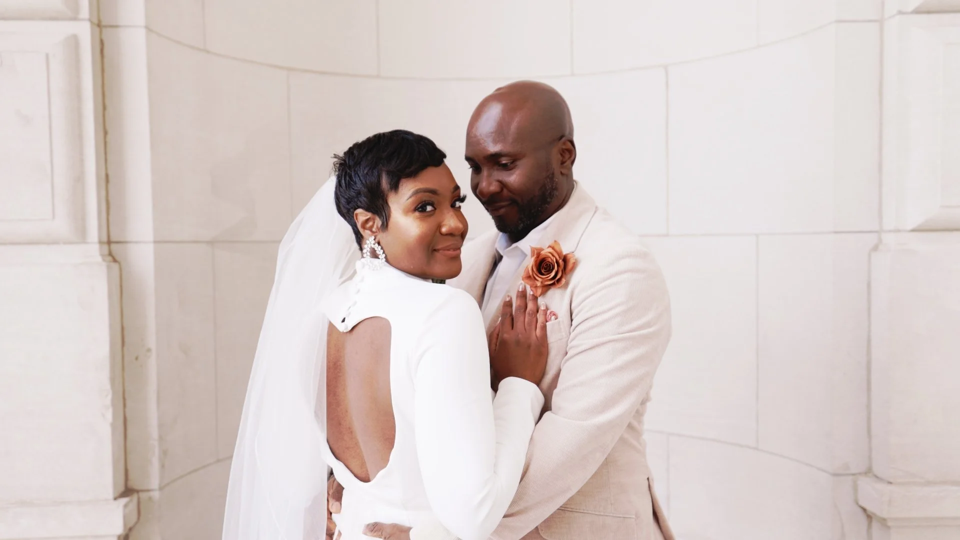 A bride and groom embracing inside a church with white stone walls, dressed in wedding attire, looking content and happy.