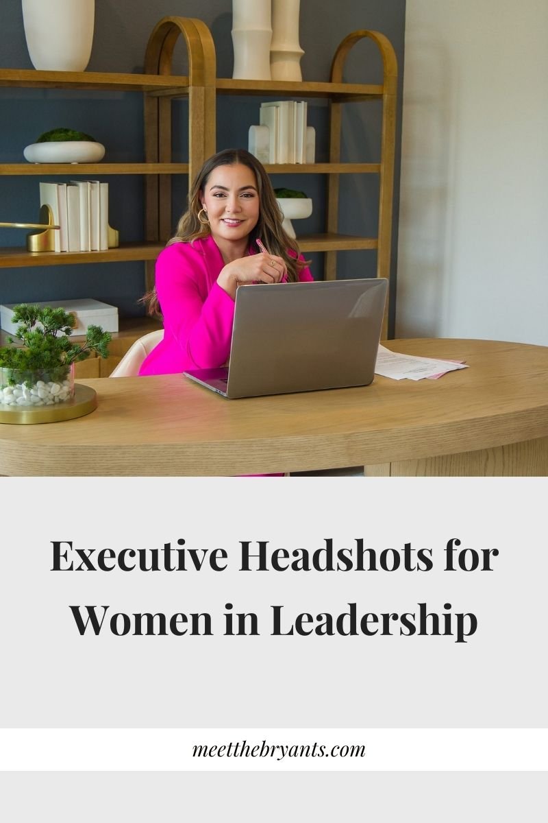 Professional woman in a bright pink suit working on a laptop at a modern wooden desk with shelving in a stylish office setting.