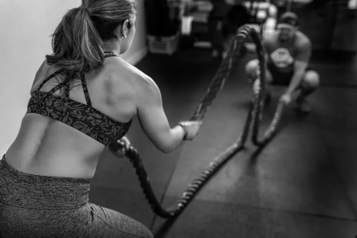 A woman with a ponytail wearing a sports bra and leggings using battle ropes for exercise, with a man in the background assisting or coaching.