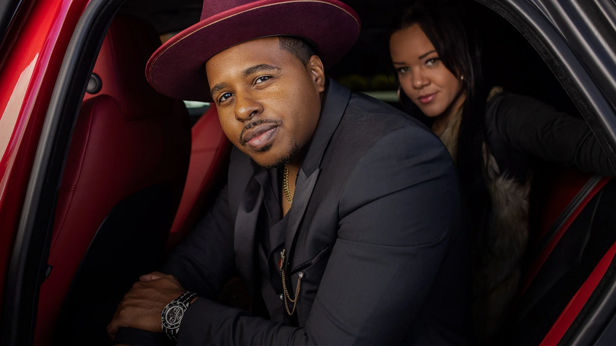 Stylish branding portrait of a man in a black suit and burgundy hat sitting in the back of a luxury car with red leather interior.